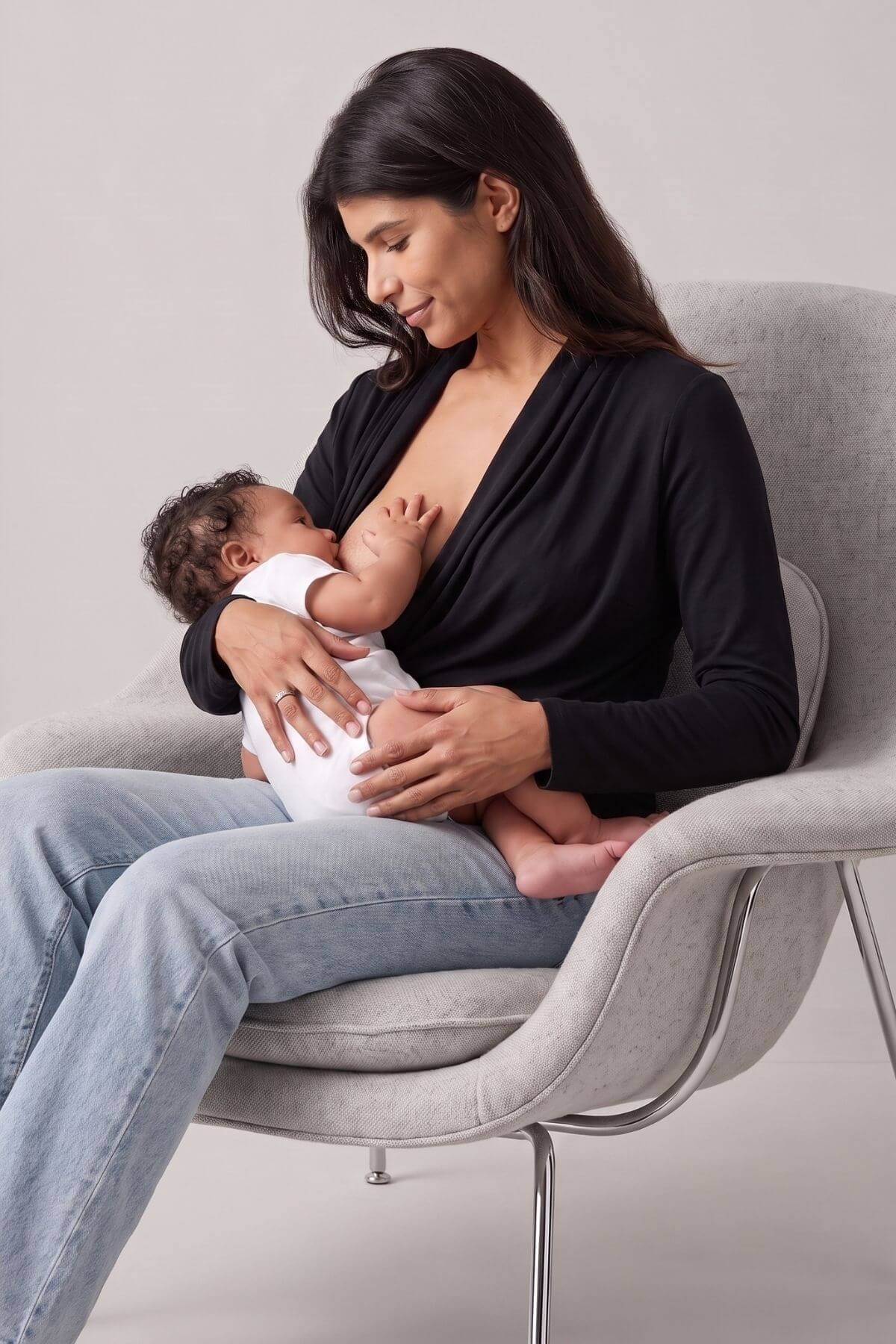 A woman in a black long-sleeve top and light blue jeans sits in a light gray chair, breastfeeding a baby dressed in a white onesie. The background is a clean, neutral studio setting.d.