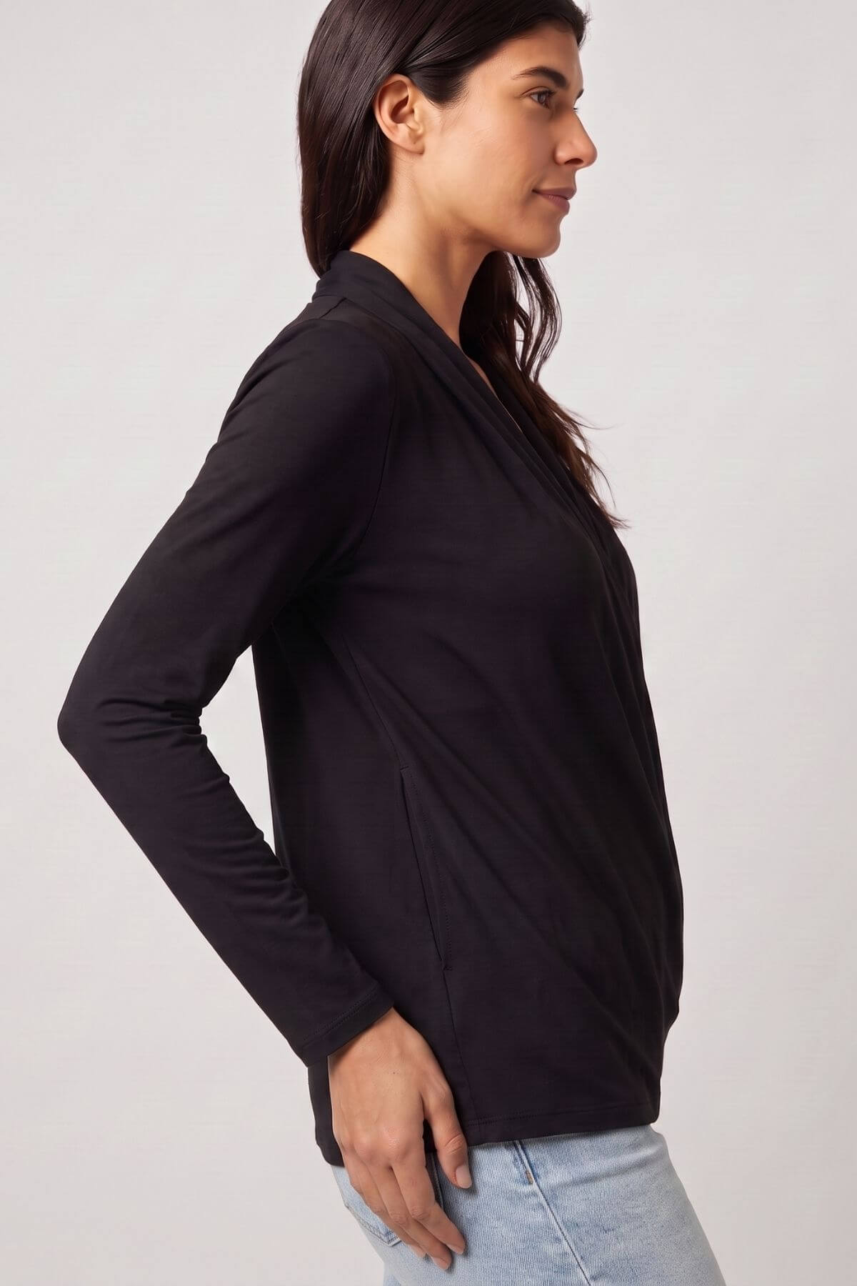 A woman wearing a black long-sleeve wrap-style top, shown in profile against a light gray studio background.d.