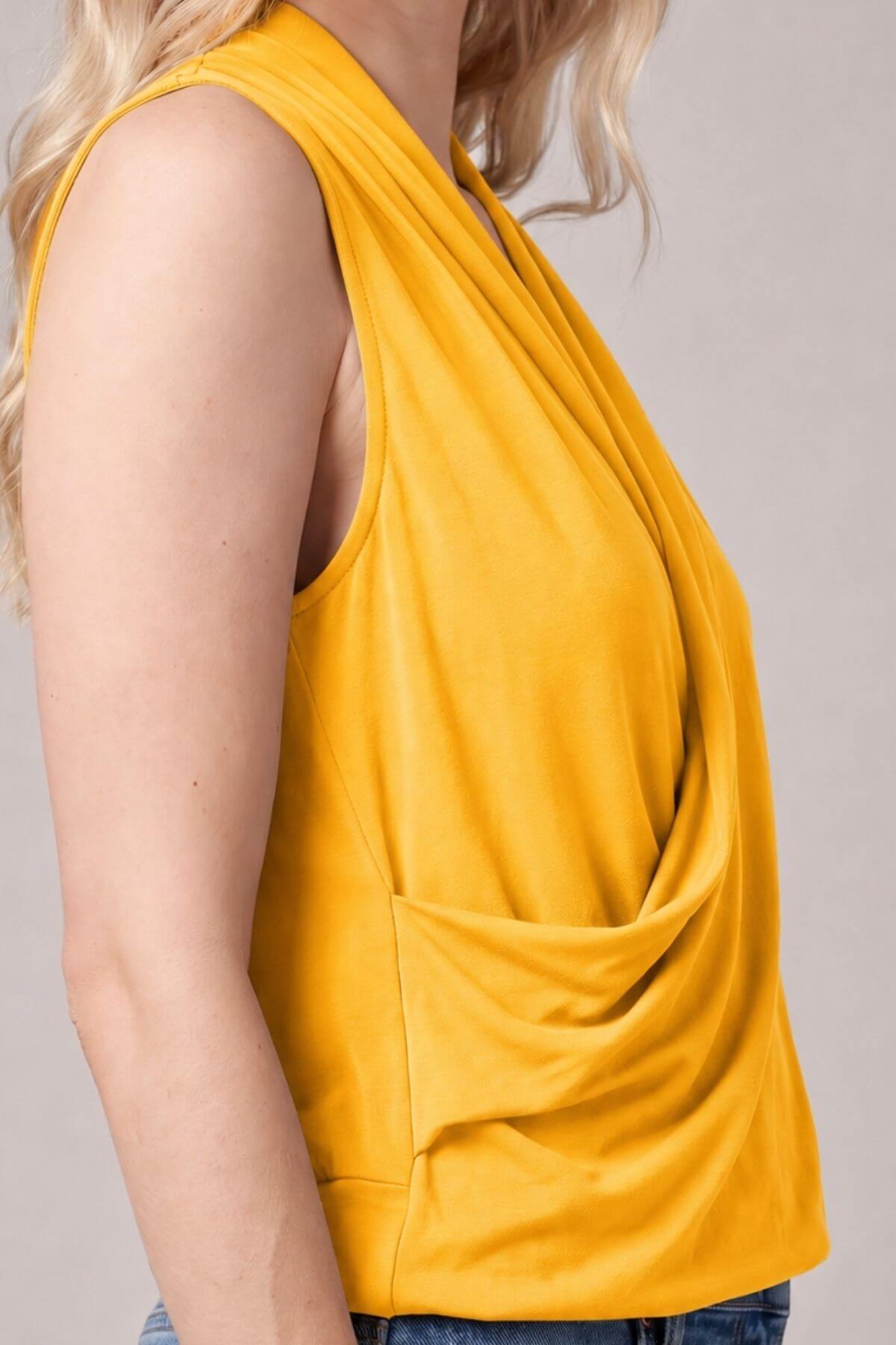 A close-up side view of a woman wearing a sleeveless draped top in a rich mustard yellow color, paired with blue jeans against a neutral gray background..