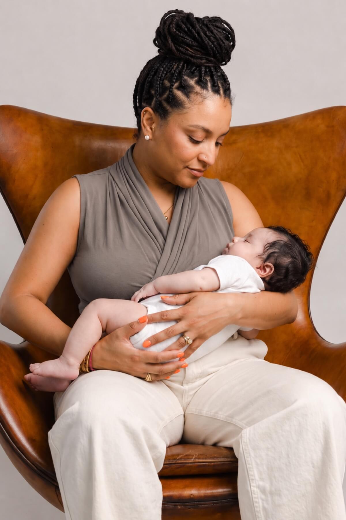 A woman in a taupe wrap top and cream pants sits in a brown leather chair, holding a sleeping newborn baby dressed in a white outfit. The background is a clean, neutral light gray studio backdrop.