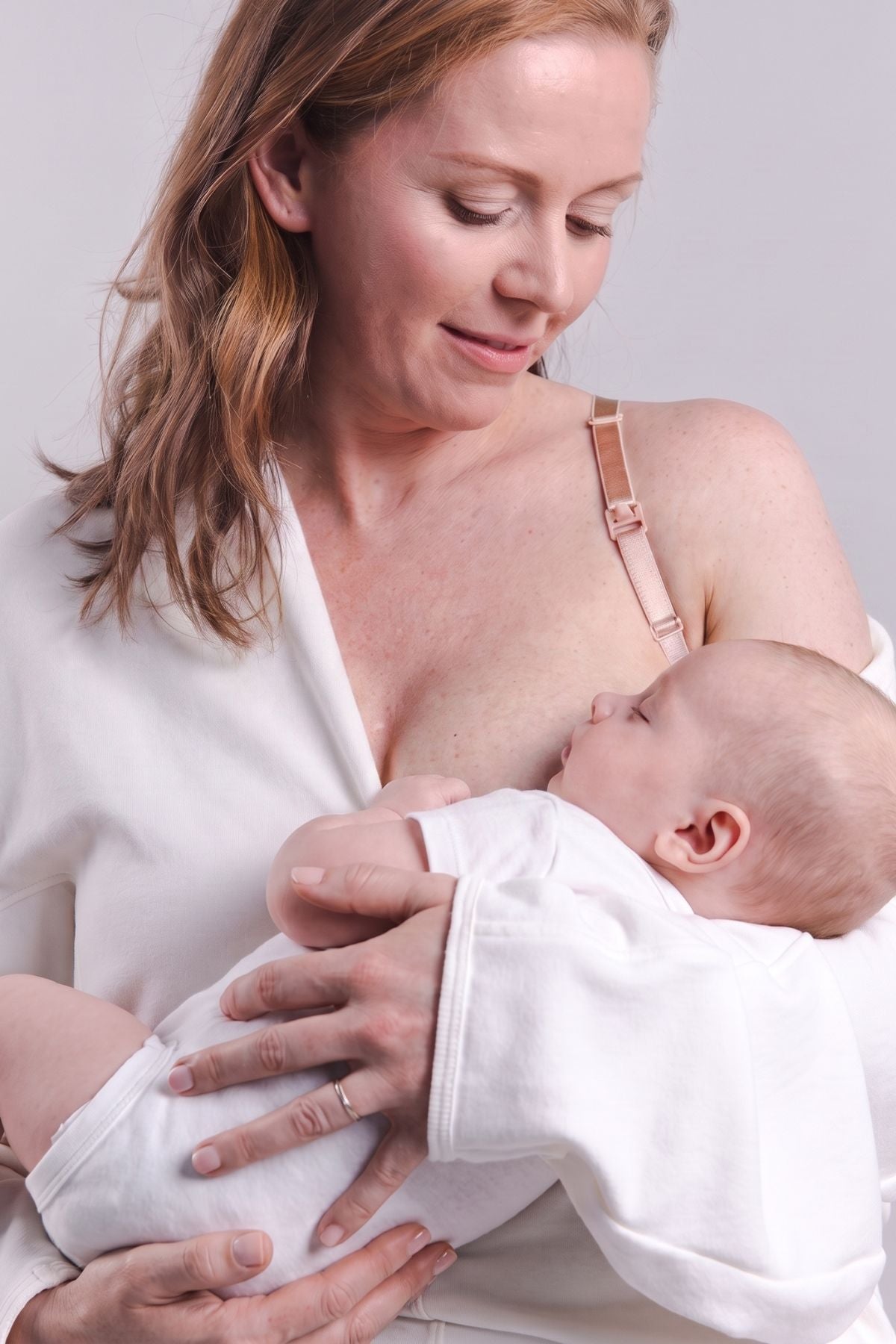 A woman wearing a white nursing top and a baby wearing a white onesie, with the woman wearing a light pink nursing bra strap visible. The woman is holding and breastfeeding the baby against a plain light gray background.
