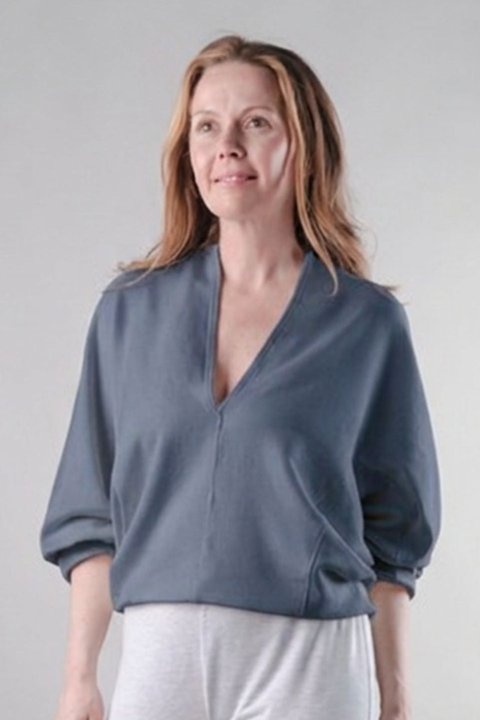 A woman modeling a slate blue V-neck blouse with three-quarter sleeves and light gray sweatpants, standing against a neutral light gray studio background.