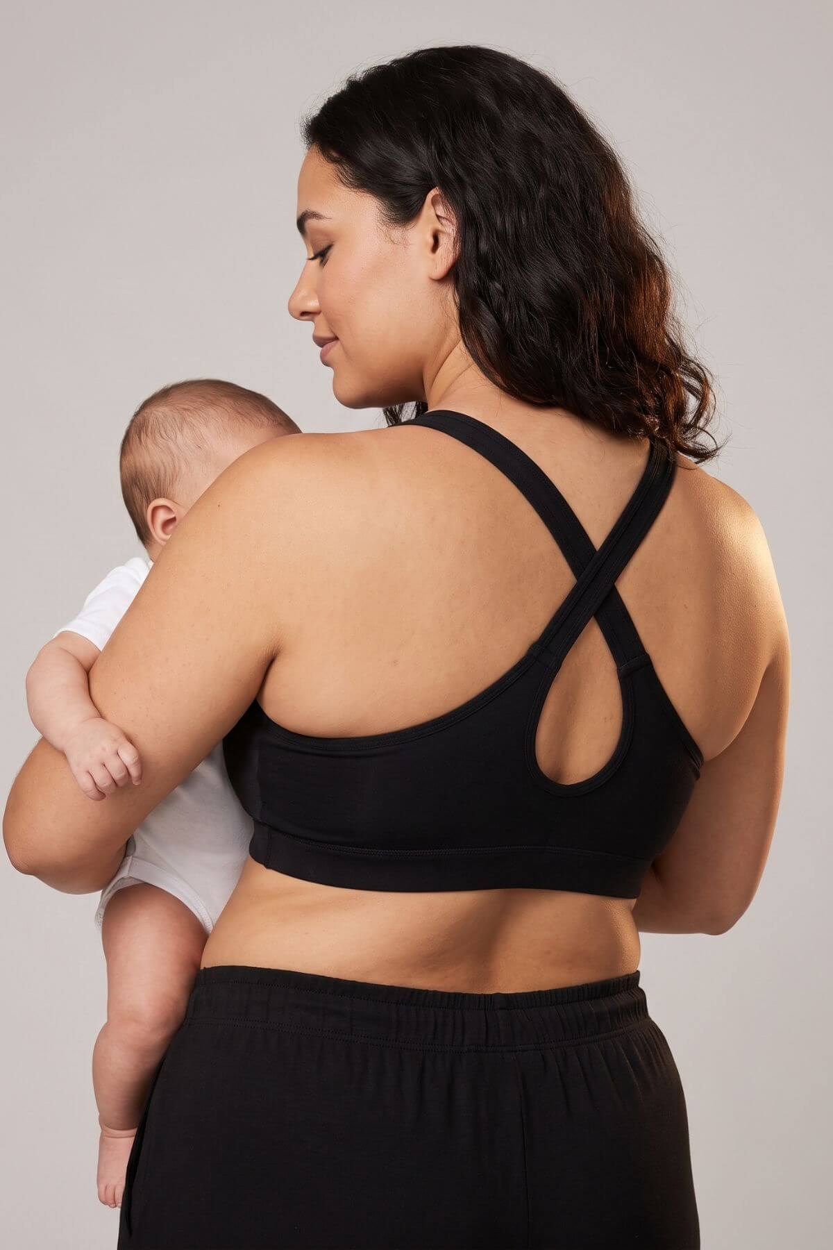 A woman wearing a black sports bra with crisscross back straps and a back cutout, holding a baby against her chest. The photo is taken from behind, showing the fit of the black bra and matching black bottoms, set against a plain light gray studio background.