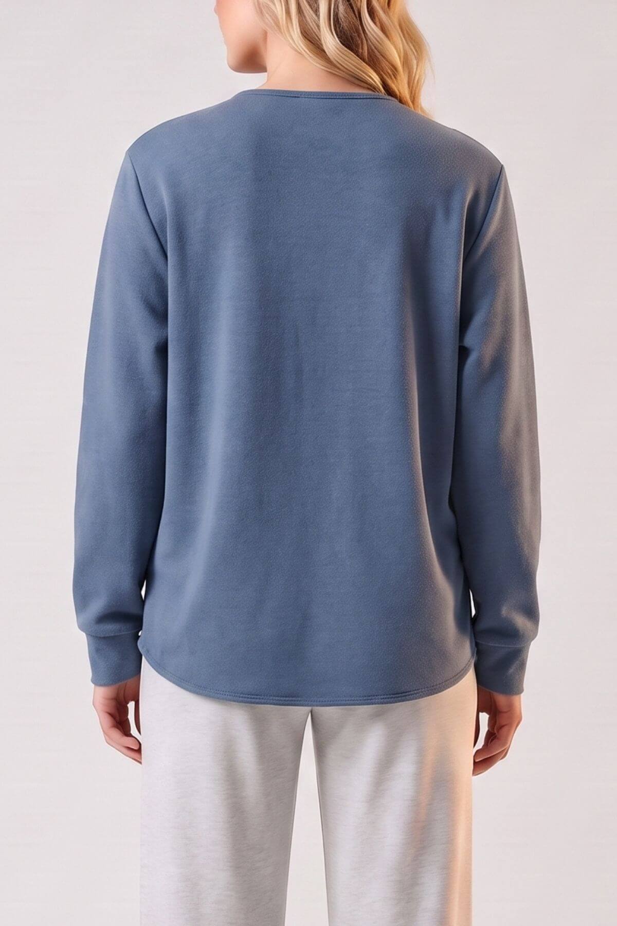 A woman shown from the back wearing a dusty slate blue long-sleeve top and light gray sweatpants against a plain light background.