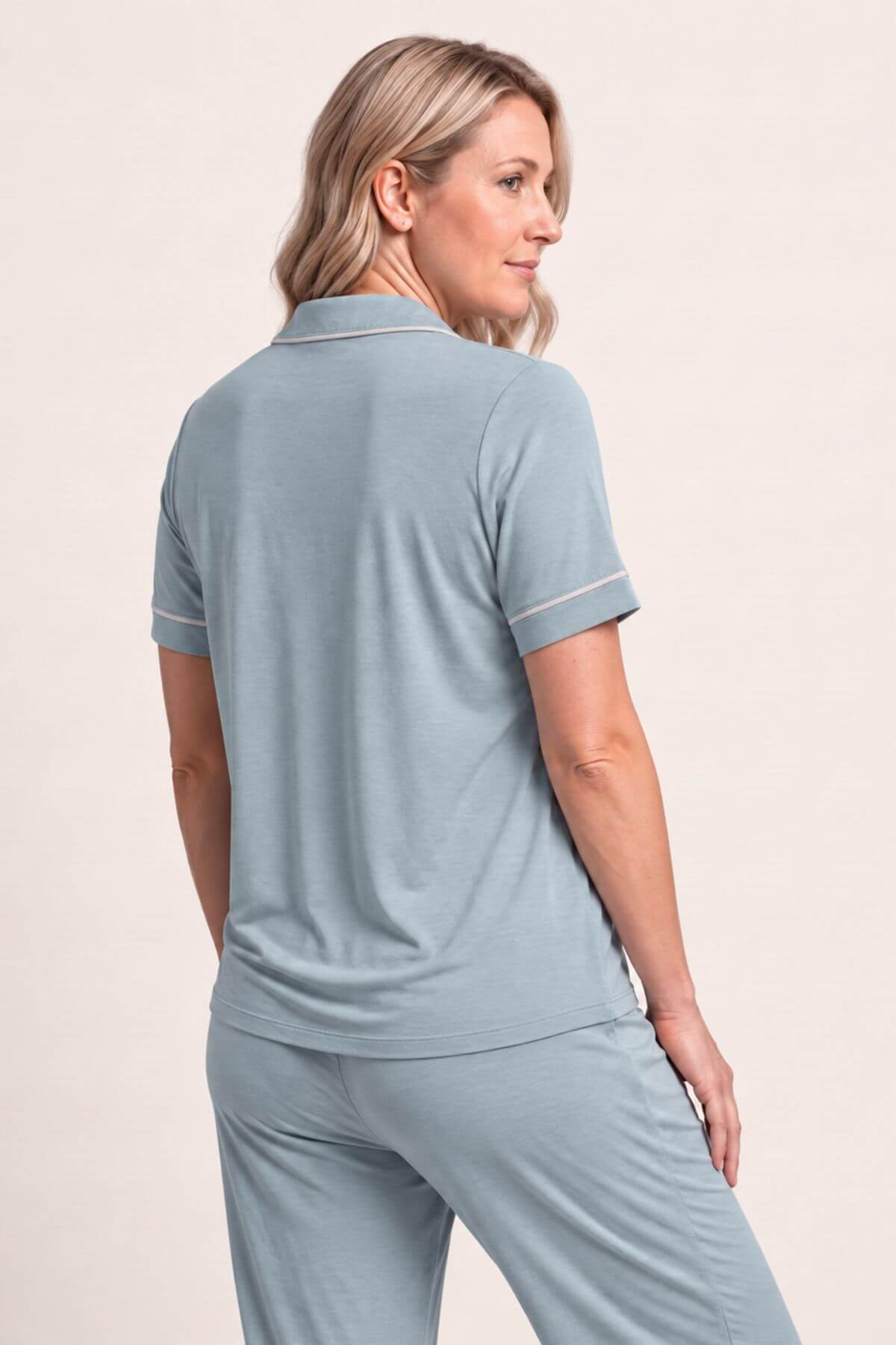 A woman wearing a light blue matching lounge set, featuring a short-sleeve polo shirt and trousers, shown from a three-quarters back view against a plain beige studio background.