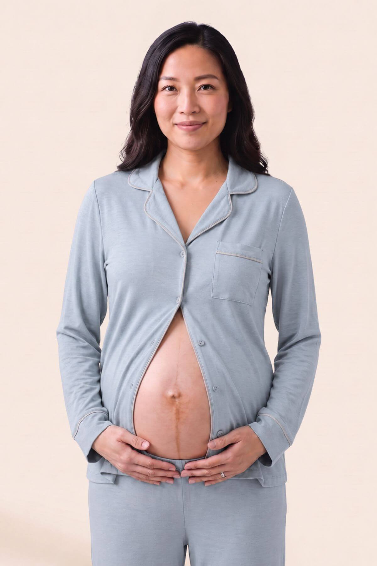 A pregnant woman wearing a light blue long-sleeve pajama top and matching pants, standing against a solid beige studio background.