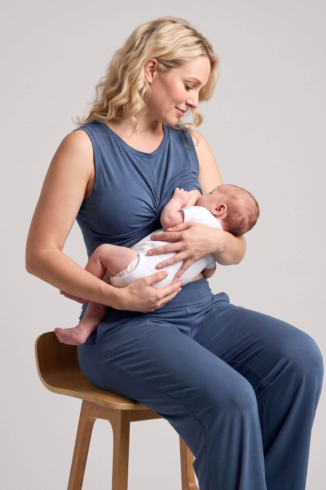 A woman in a navy blue nursing set sits on a wooden stool, breastfeeding a baby dressed in a white outfit against a neutral light gray background.