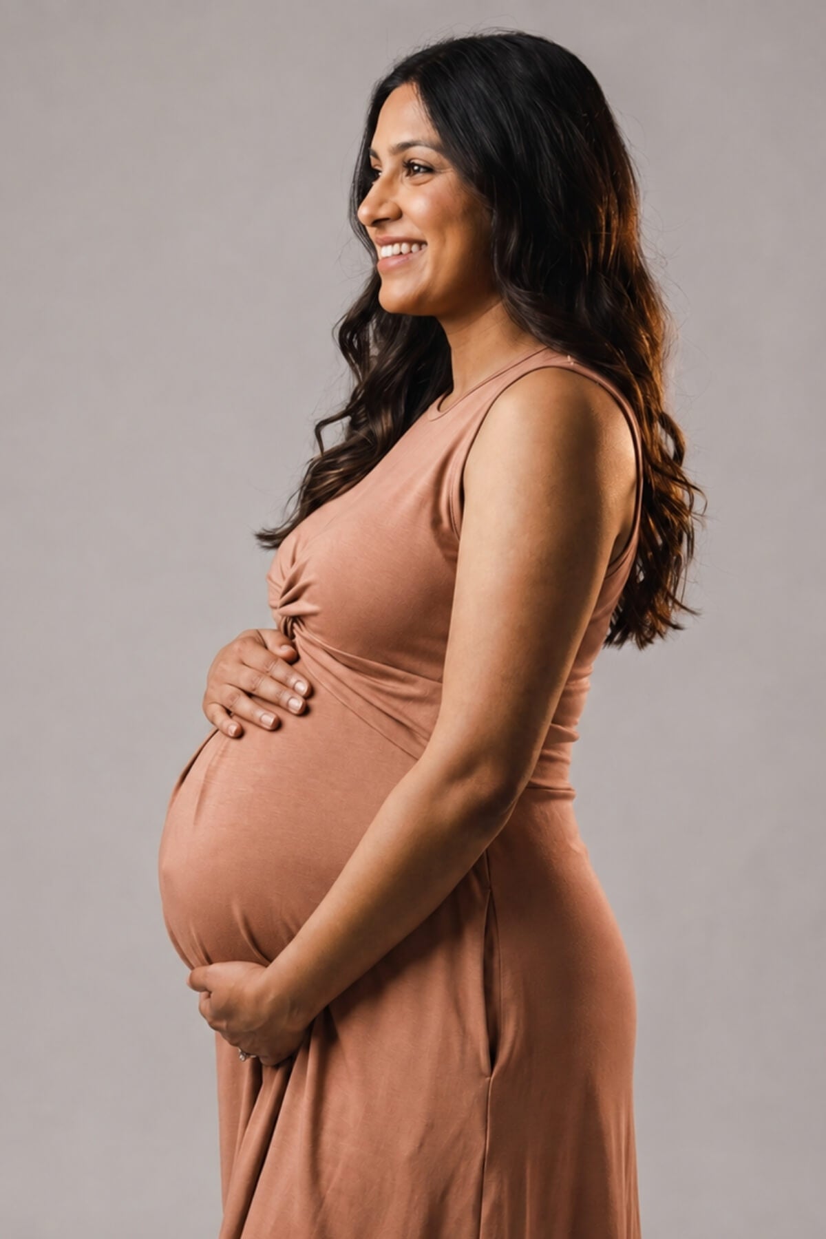A smiling pregnant woman in profile wearing a sleeveless dusty rose maternity dress, holding her baby bump against a solid gray studio background.
