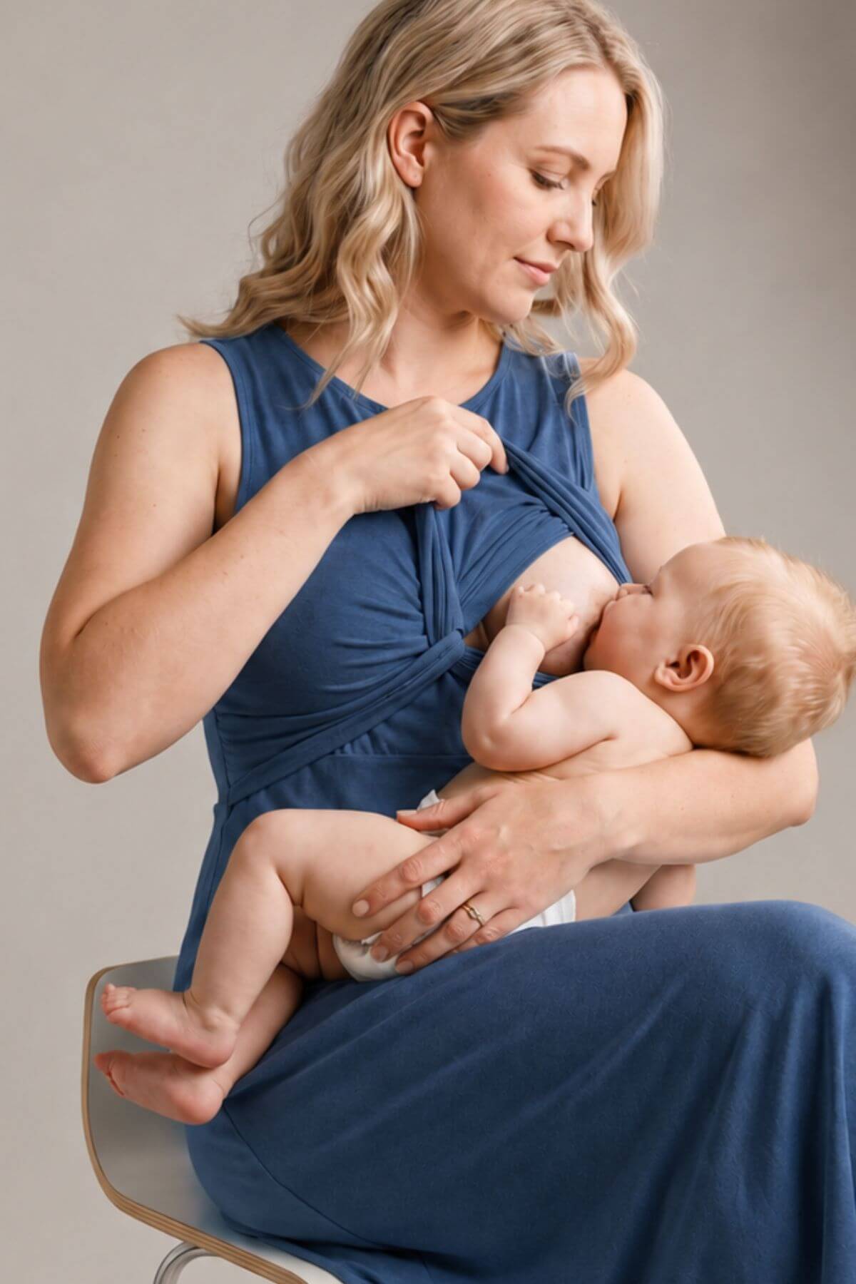A woman wearing a blue nursing tank top and matching pants holds a baby while breastfeeding, seated on a chair against a neutral gray background.