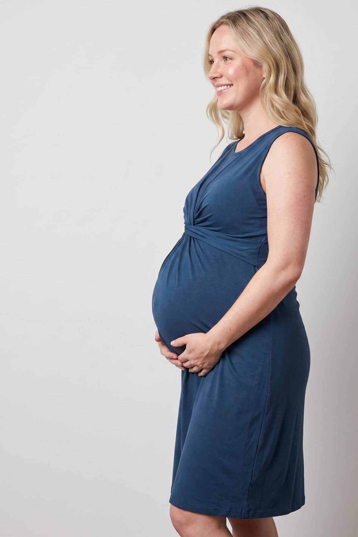 A pregnant woman in side profile wearing a sleeveless, slate blue maternity dress, standing against a neutral light gray studio background.