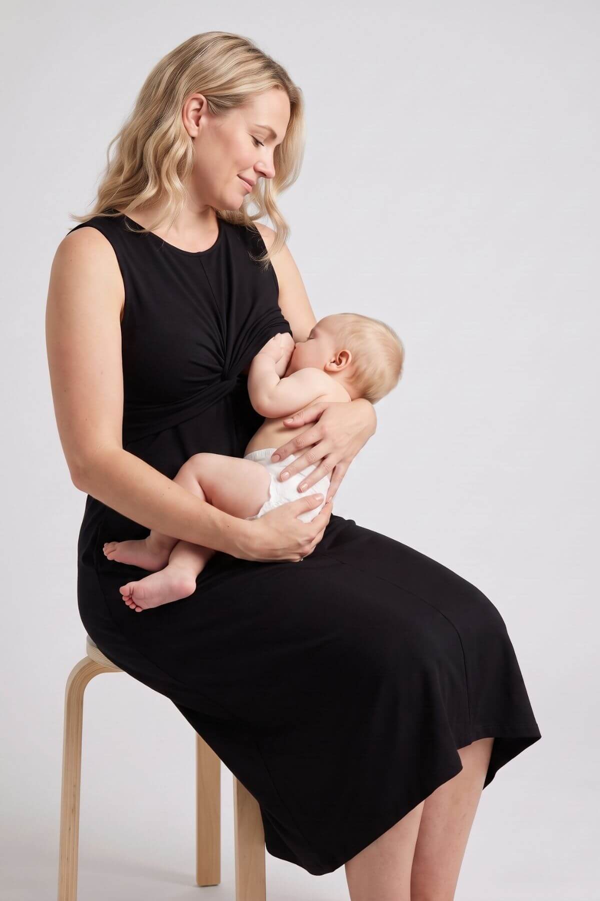 A woman in a black sleeveless dress sits on a wooden chair, breastfeeding a baby wearing a white diaper against a light gray studio background..