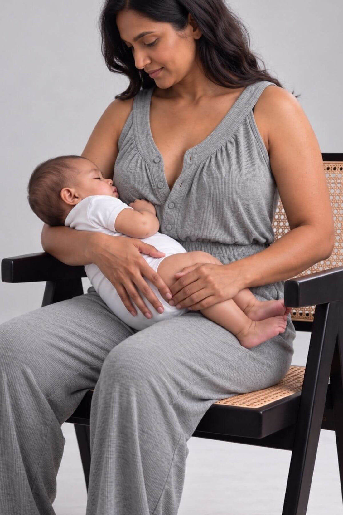 A woman wearing a gray sleeveless baby nursing top and matching pants sits on a black chair with a woven seat, holding a sleeping baby dressed in a white onesie.