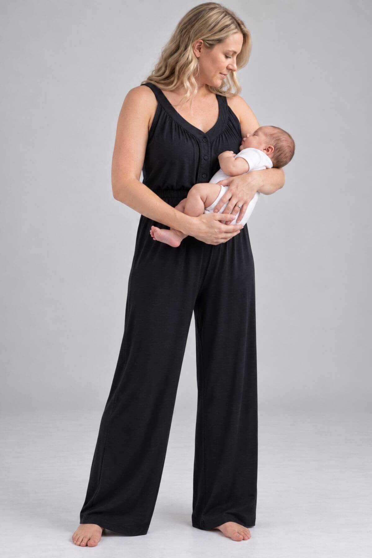 A woman wearing a black sleeveless jumpsuit with wide legs holds a sleeping newborn baby dressed in a white outfit, standing against a neutral light gray studio background.