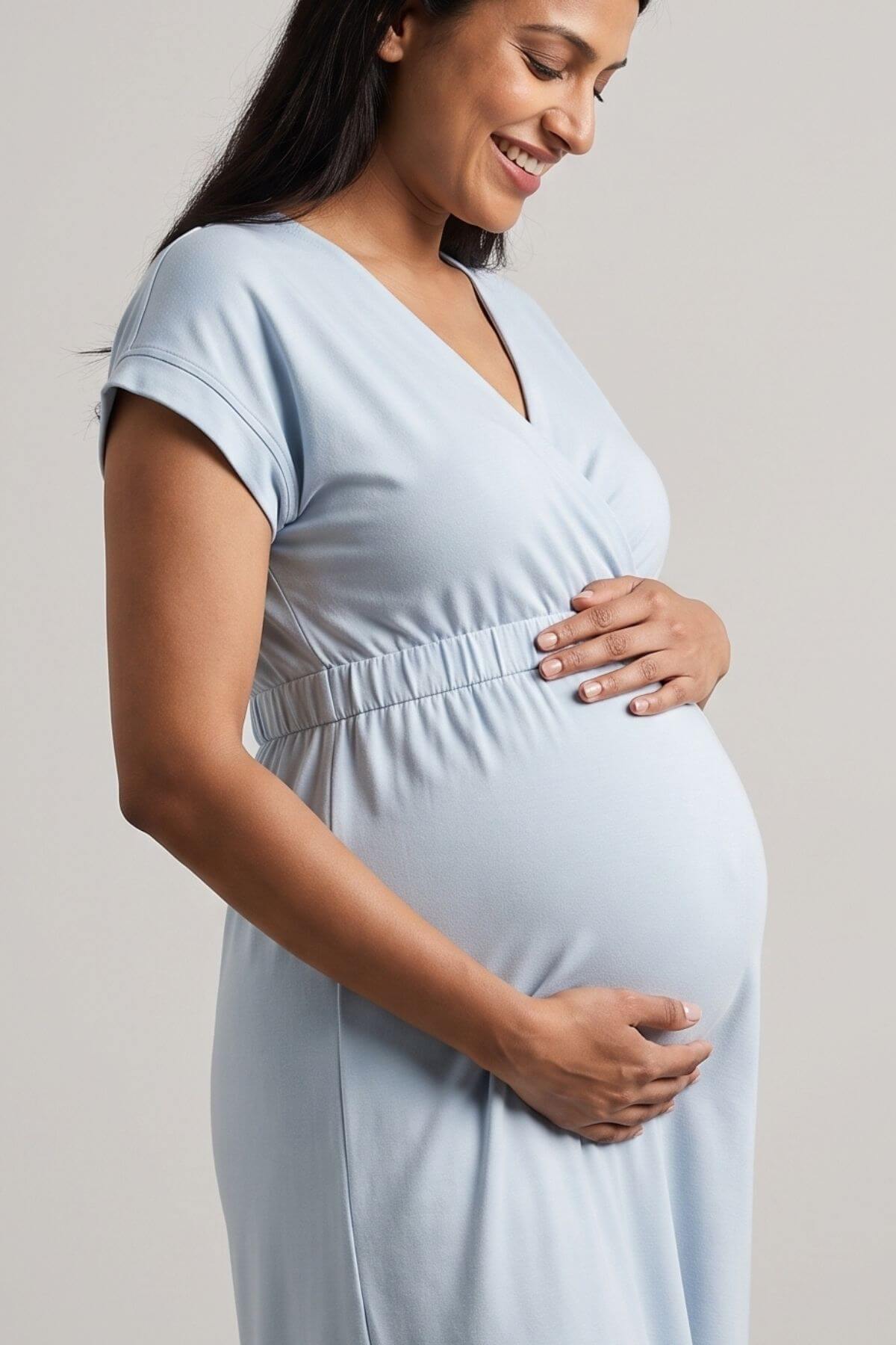 A pregnant woman wearing a light blue maternity dress, holding her baby bump against a neutral light gray background.