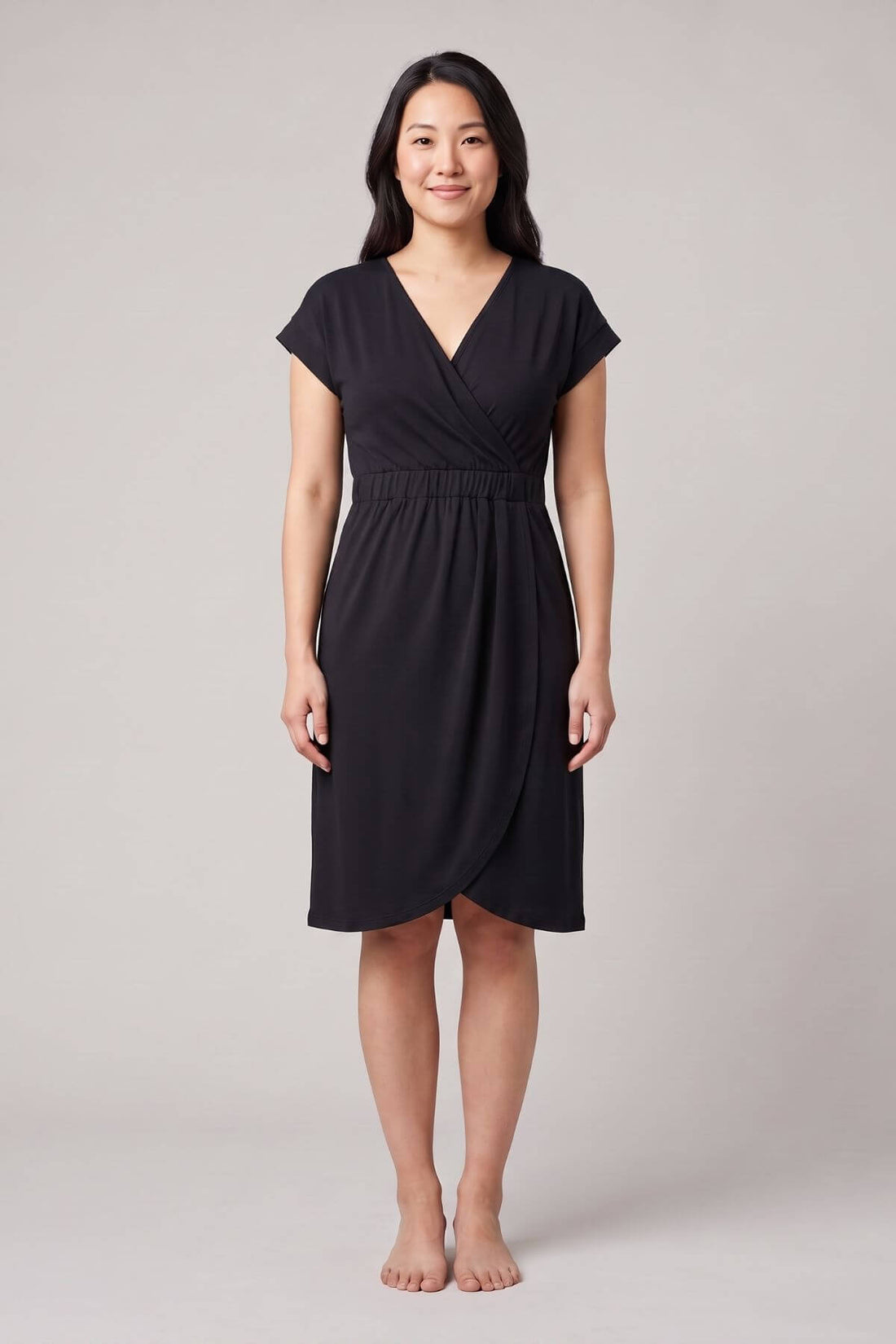 A woman modeling a black short-sleeve wrap-style dress with a V-neckline and gathered waist, standing against a plain light gray studio background.