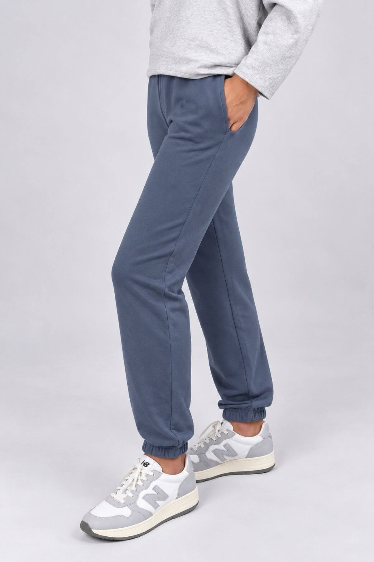 A person wearing slate blue jogger pants with elastic cuffs, paired with a light gray sweatshirt and gray New Balance sneakers, shown from the waist down against a light gray studio background.