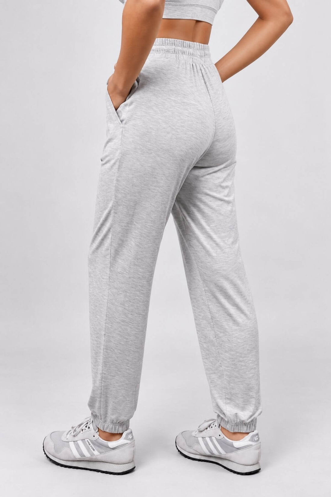 A person wearing light heather gray jogger sweatpants with elastic cuffs and side pockets, paired with white sneakers, shown from the back against a light gray studio background.
