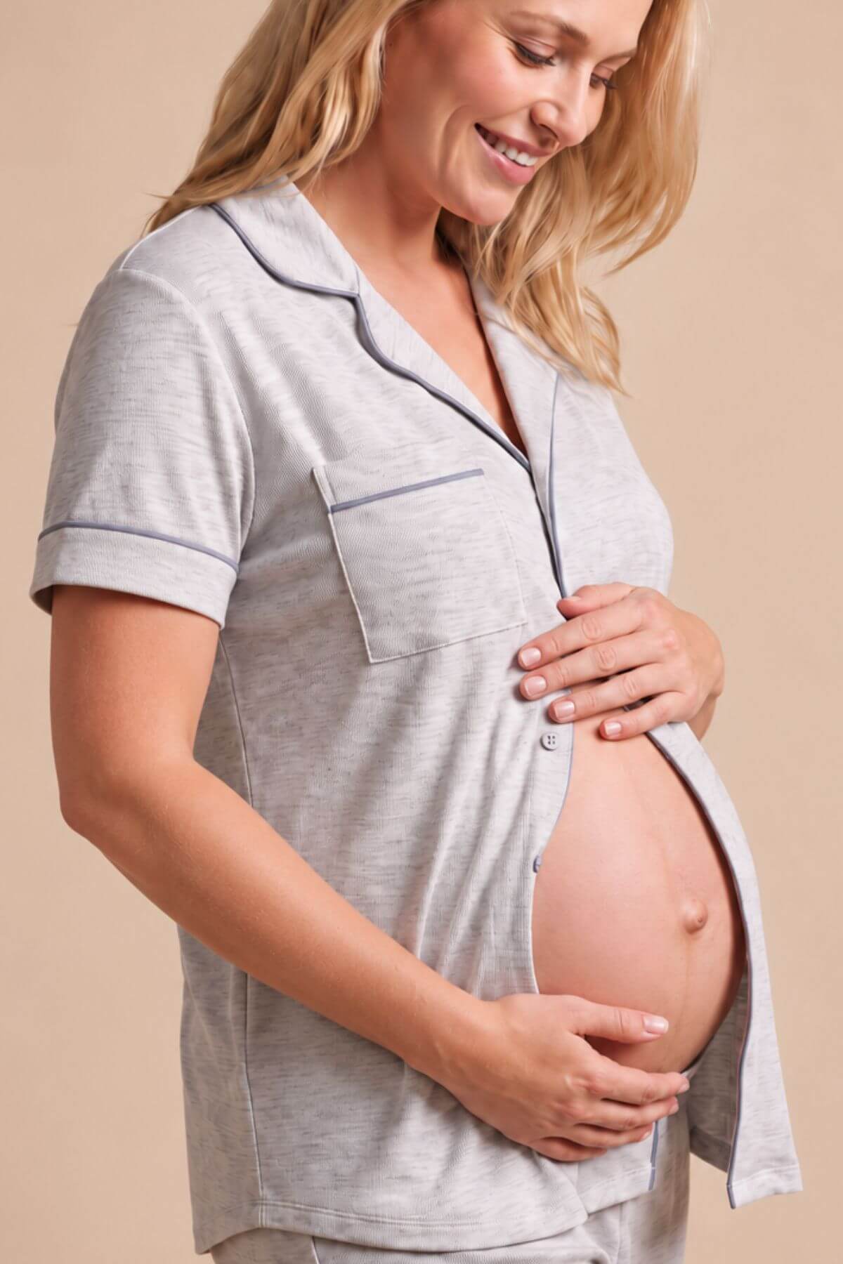 A pregnant woman wearing a light gray short-sleeve pajama top with dark gray piping, holding her baby bump against a warm beige background.