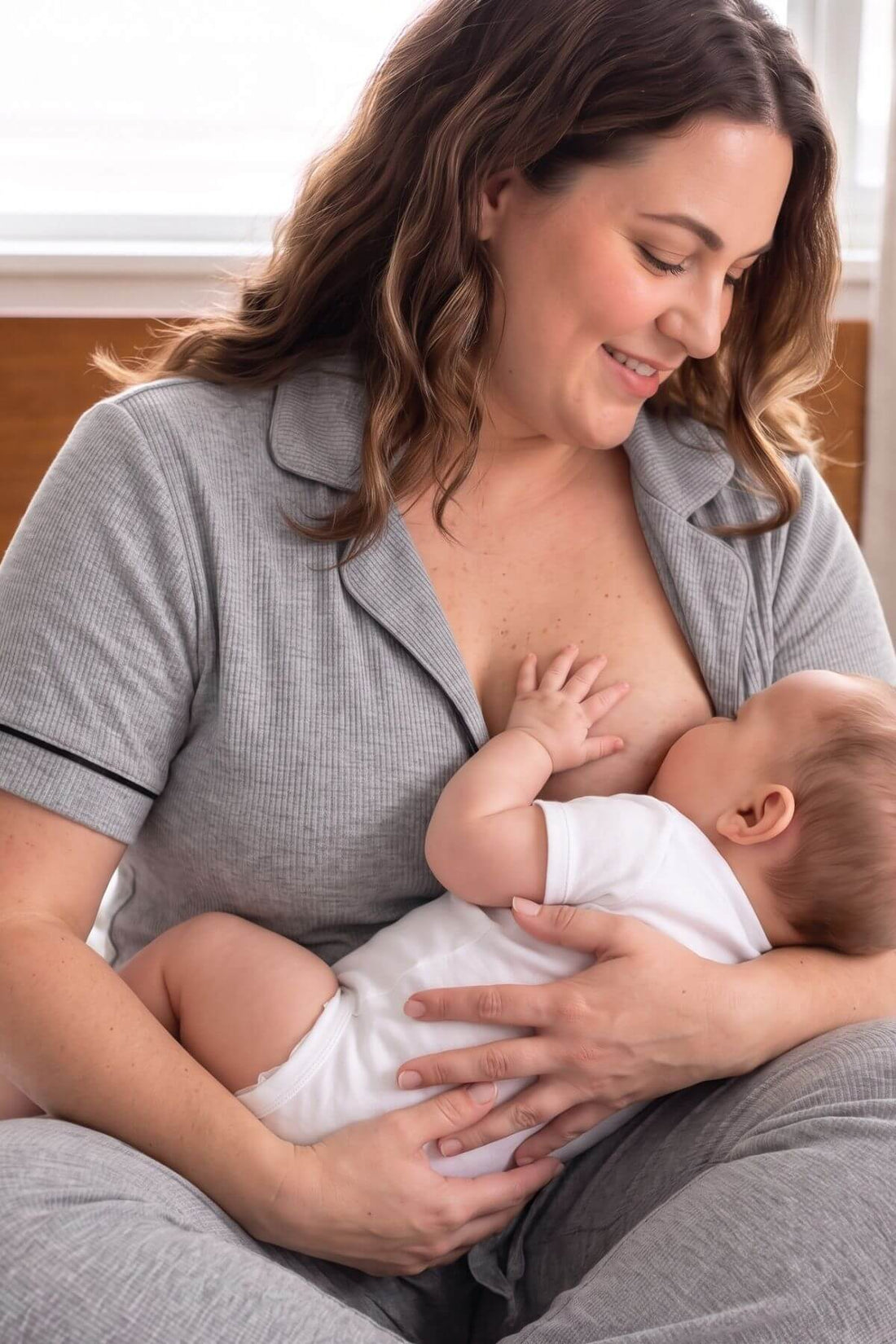 A woman in a gray short-sleeve pajama top and matching pants holds a baby wearing a white onesie while breastfeeding in a softly lit indoor .
