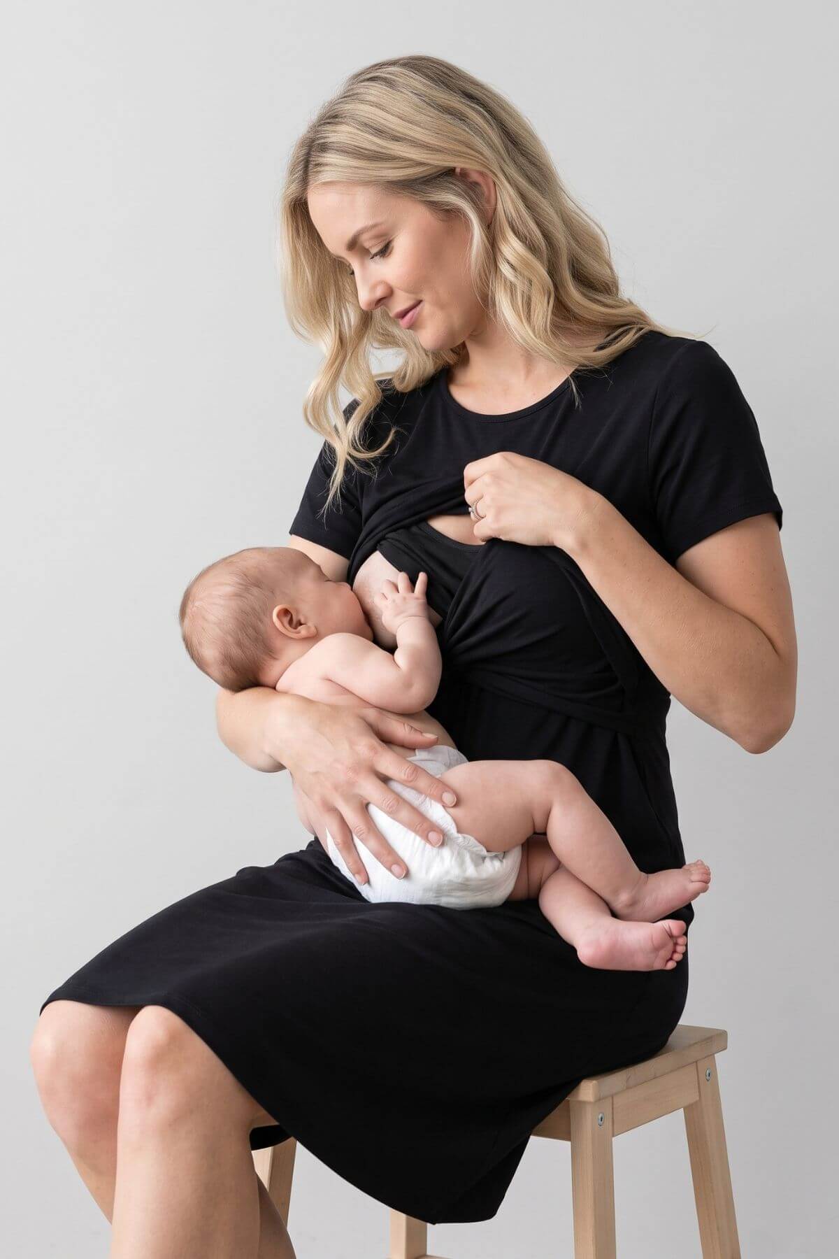 A woman in a black short-sleeve maternity dress sits on a wooden stool, breastfeeding a baby wearing a white diaper. The photo is shot in a studio setting against a light gray background..