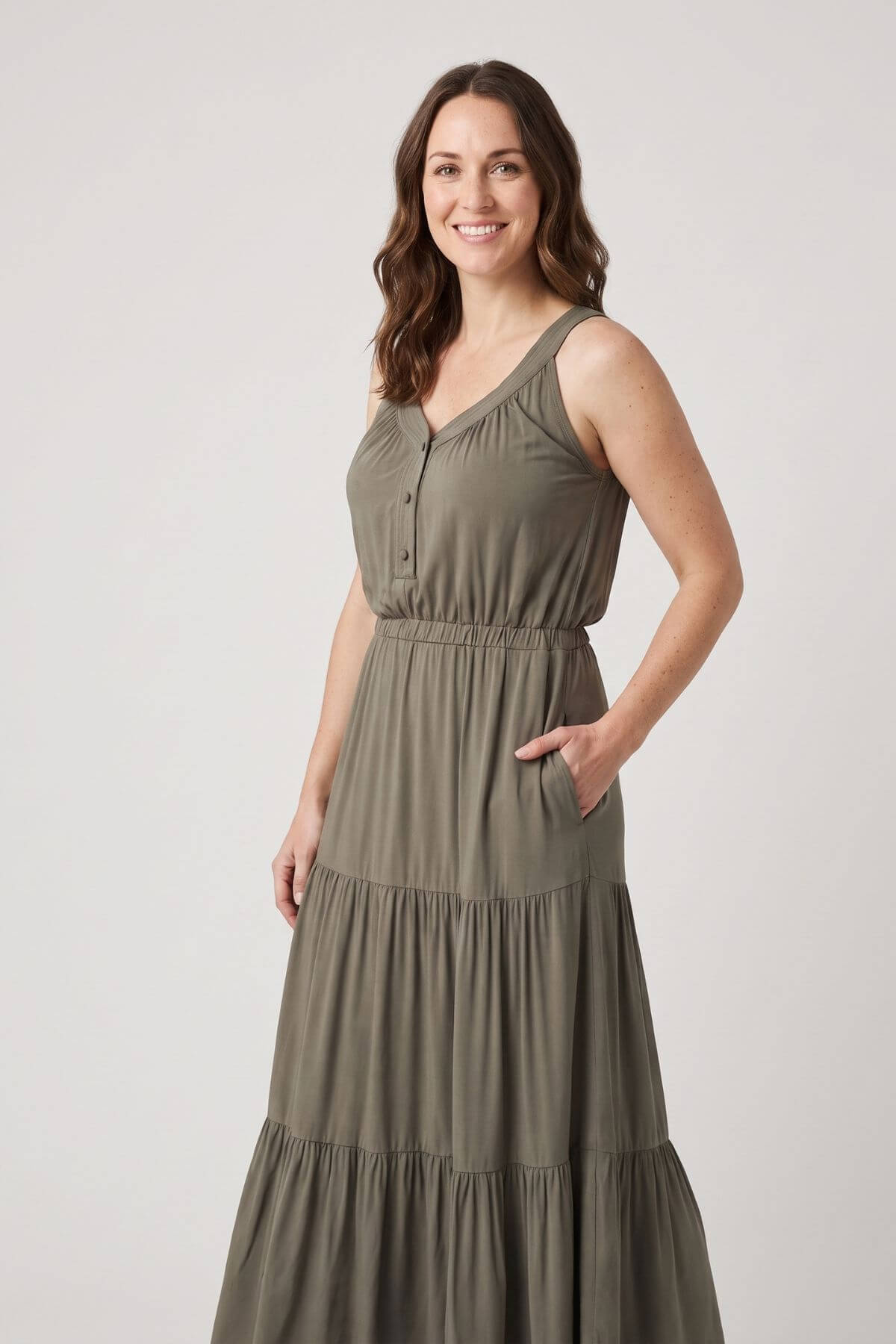 A woman modeling a sleeveless, tiered maxi dress in a muted olive green color, standing against a plain light gray studio background.