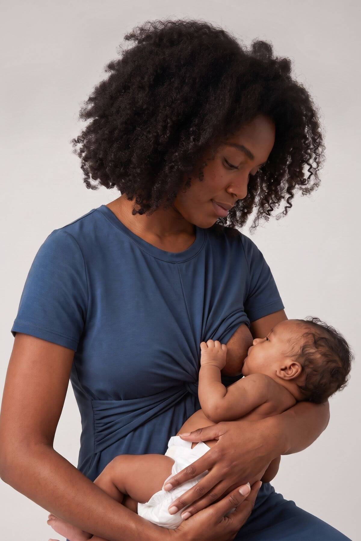 A woman with curly black hair wearing a navy blue twist-front nursing top holds and breastfeeds a baby wrapped in a white blanket, set against a plain light gray studio background.