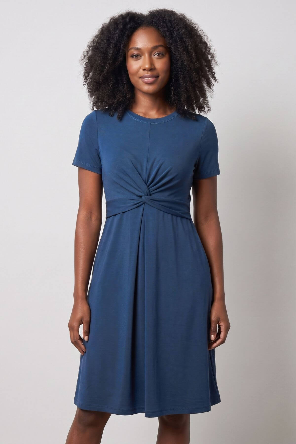 A woman with curly black hair wearing a short-sleeve, knee-length teal blue dress with a twisted knot detail at the waist, standing against a light gray studio background.