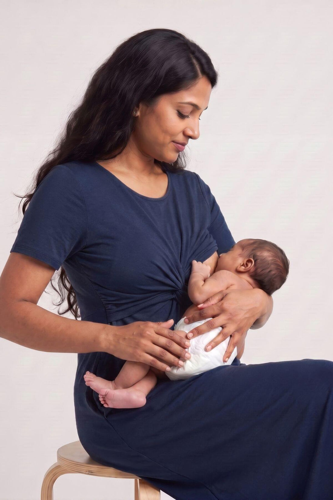 A woman wearing a navy blue maternity nursing dress sits on a wooden stool, breastfeeding a newborn baby wrapped in a white swaddle. The photo is shot against a clean, light off-white studio background.