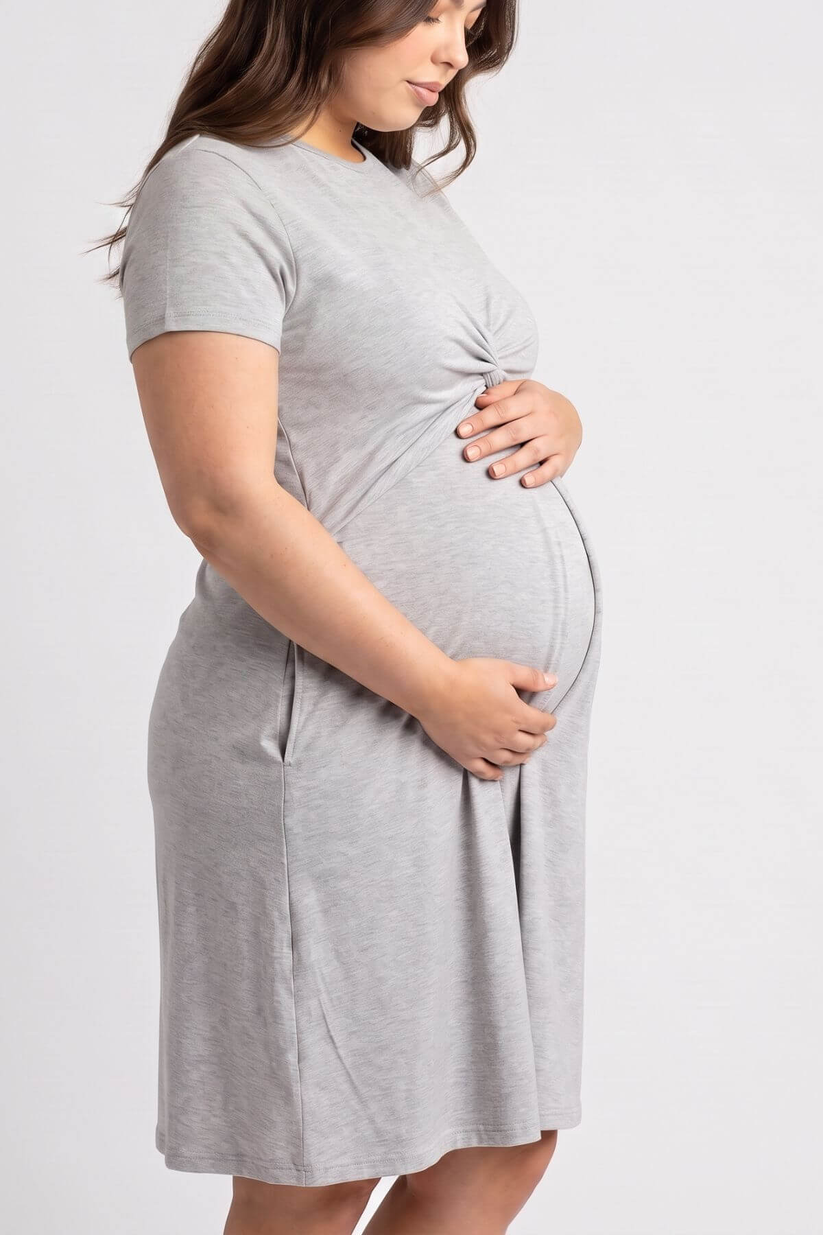 A pregnant woman wearing a light gray short-sleeve maternity dress with side pockets, standing against a plain light gray studio background.