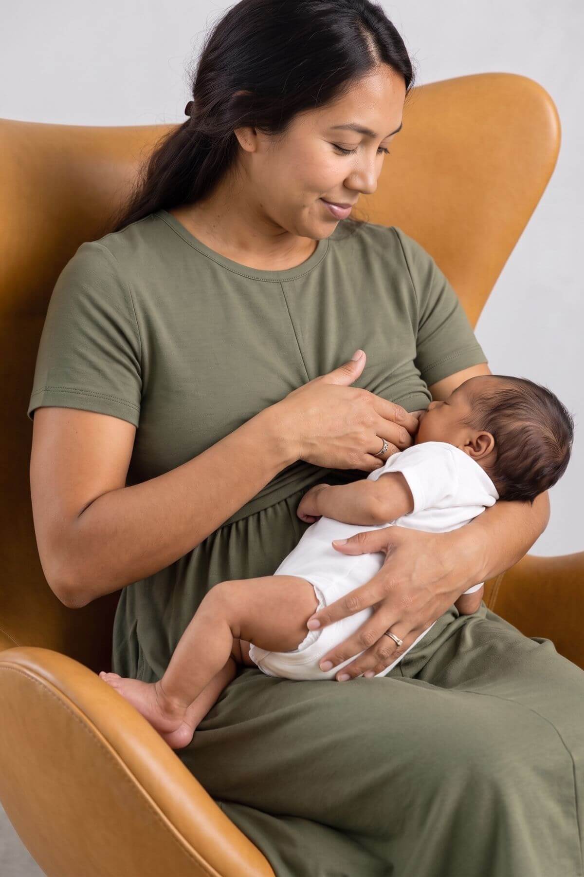 A woman in an olive green maternity dress nursing a baby wearing a white onesie while sitting in a tan leather armchair.