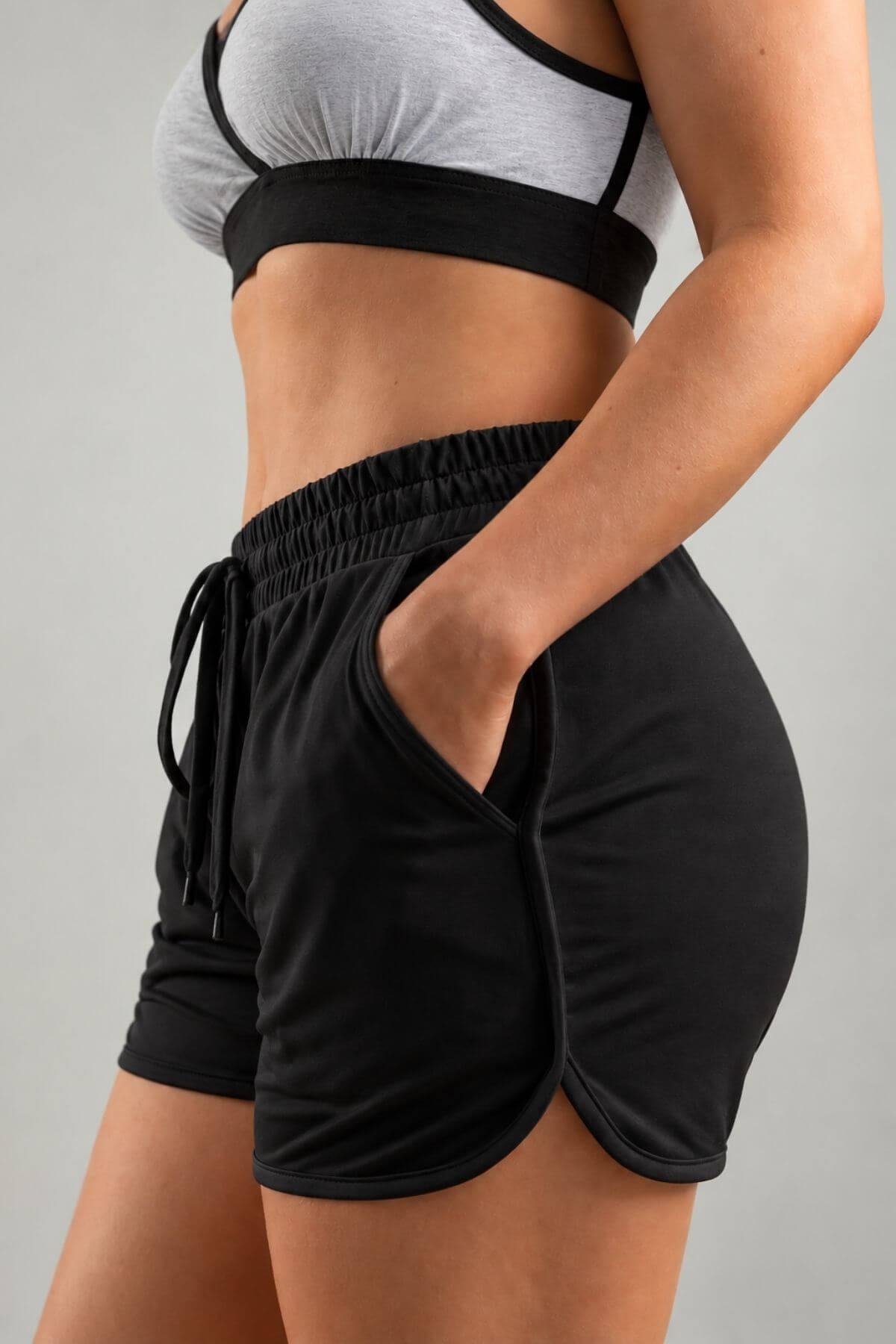 A close-up of a model wearing black athletic drawstring shorts with side pockets and a gray sports bra against a light gray background.