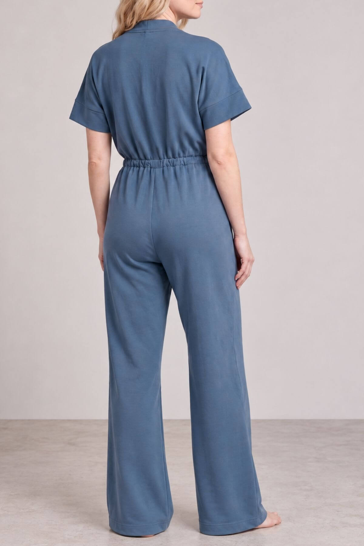 A woman viewed from the back wearing a slate blue short-sleeve jumpsuit with a high waist and wide legs, standing against a neutral studio backdrop..