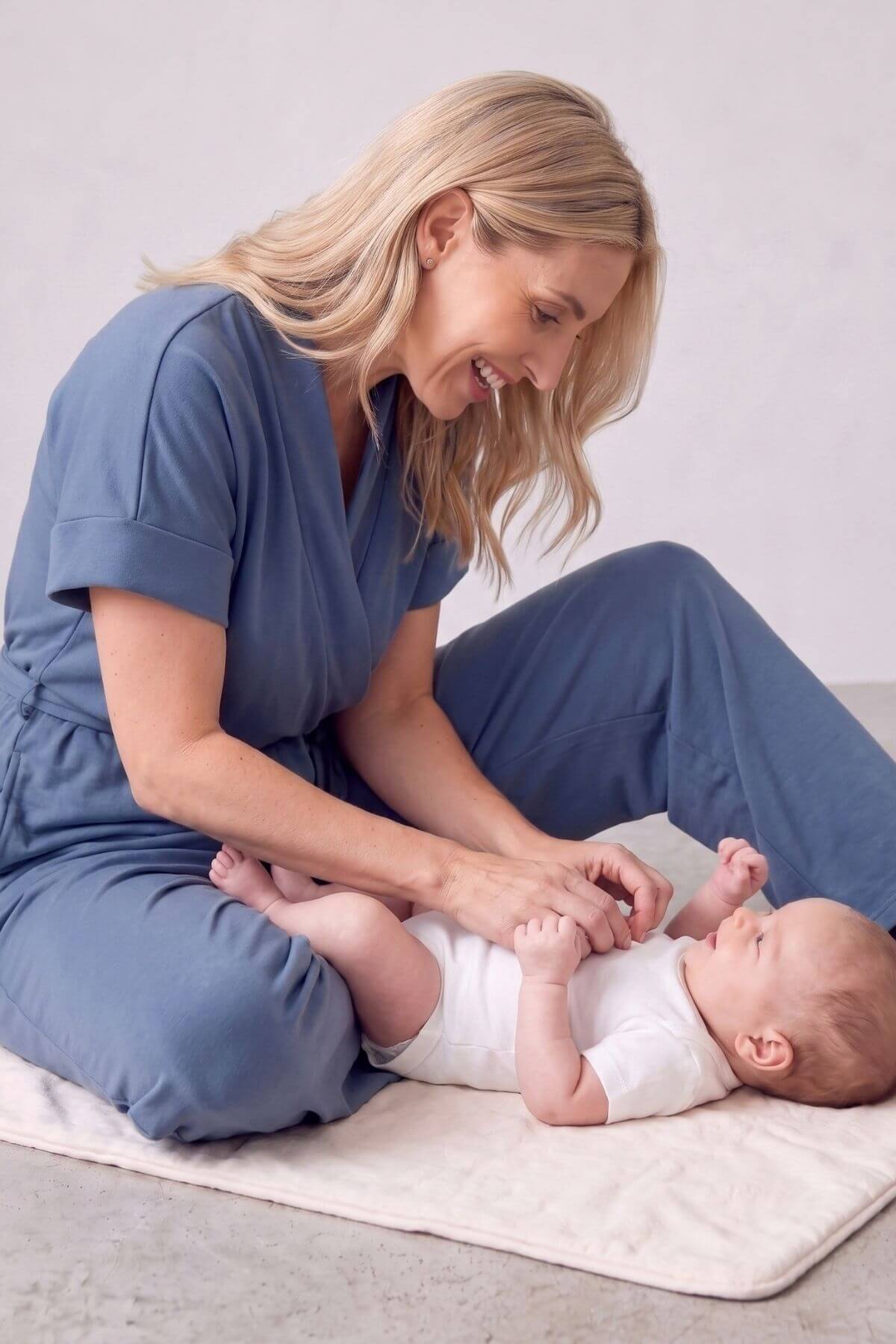 A woman in blue medical scrubs sits on a light beige mat, holding and washing a baby wearing a white onesie in a bright, clean room..