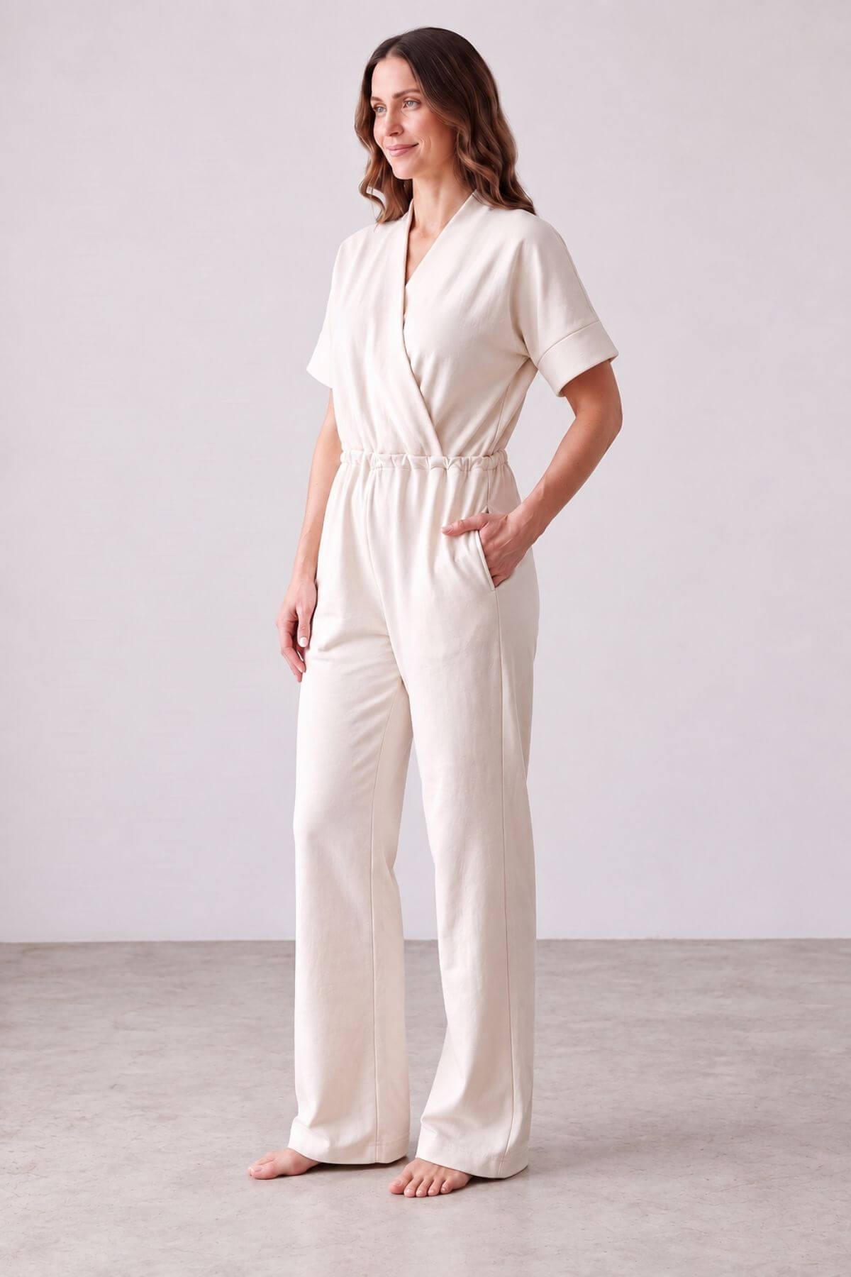 A woman modeling a short-sleeve cream-colored jumpsuit with a wrap-style V-neck and wide-leg pants, standing barefoot against a neutral studio backdrop.