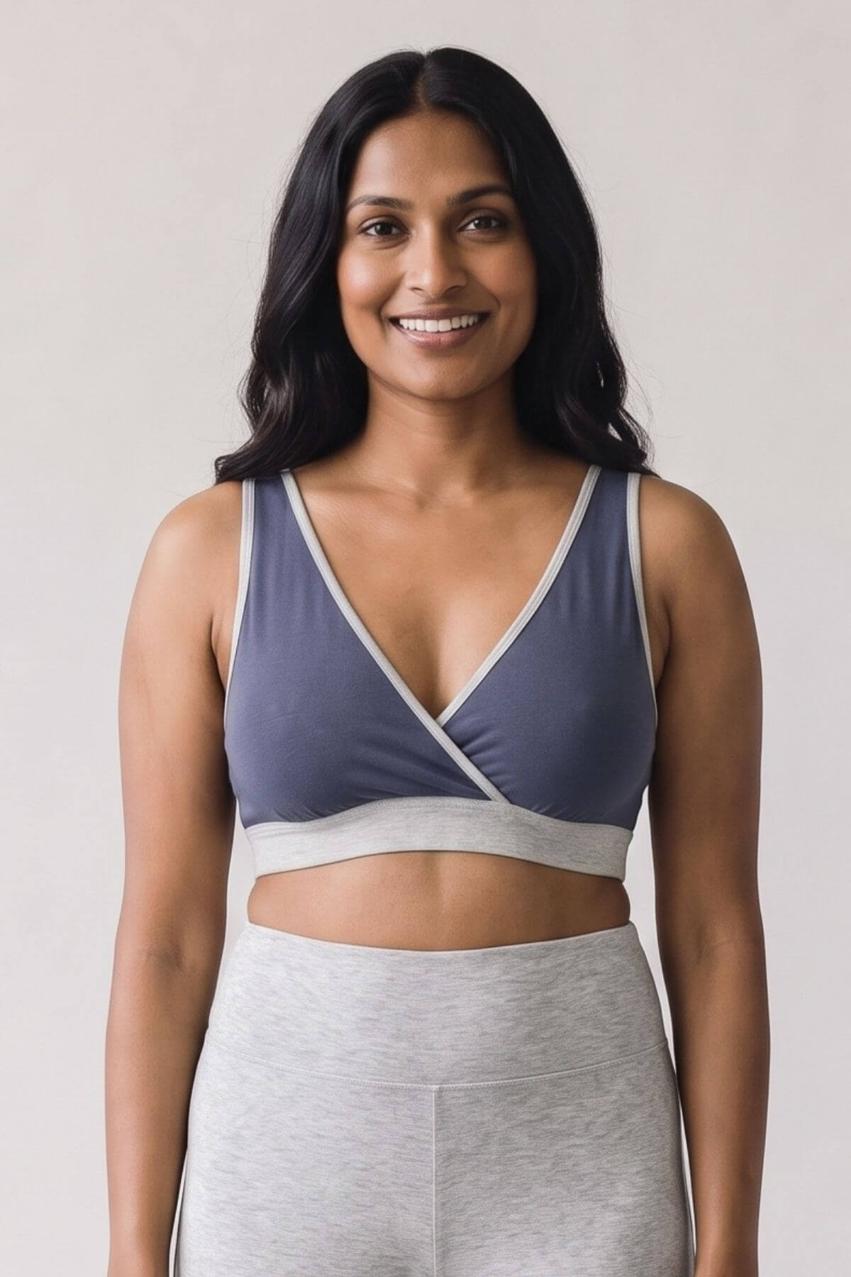 A woman modeling a slate blue and light gray wrap-style sports bra with matching gray athletic leggings, photographed against a plain light gray studio background.