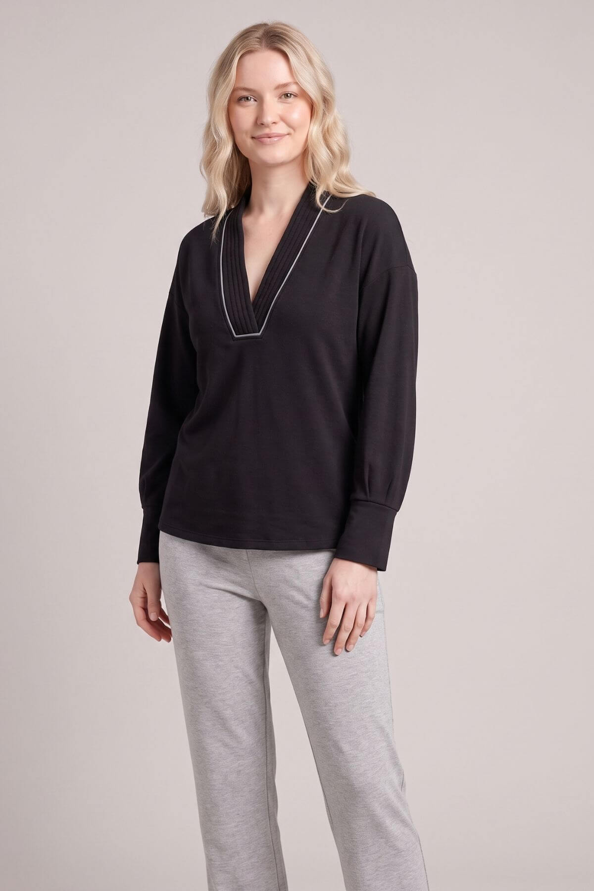 A woman modeling a black long-sleeve top with a V-neck and white piping trim, paired with light gray sweatpants, against a solid light gray studio background.