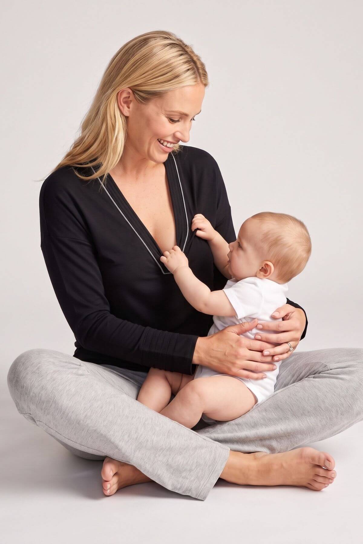 A woman sitting cross-legged on a light gray studio floor holds a baby. She wears a black long-sleeve V-neck top and light gray sweatpants, and the baby is dressed in a white outfit..