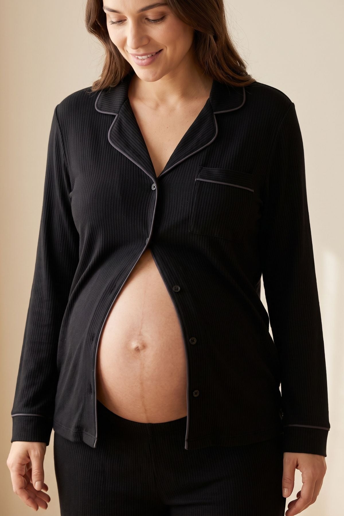 A pregnant woman wearing a black ribbed long-sleeve pajama top with dark piping and matching pants, shown against a warm beige background.