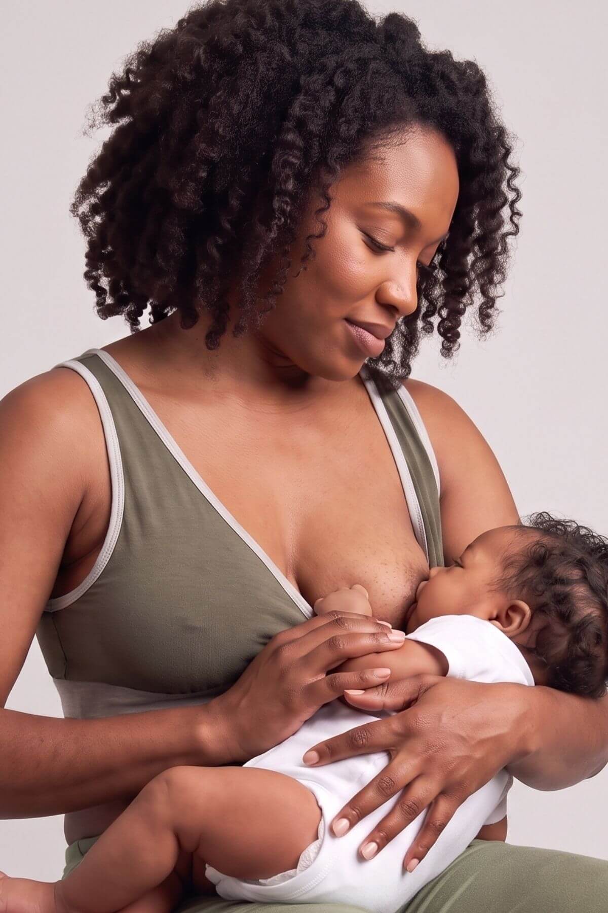 A woman with curly dark hair breastfeeding a baby, wearing an olive green nursing tank top and matching bottoms against a plain light gray background.