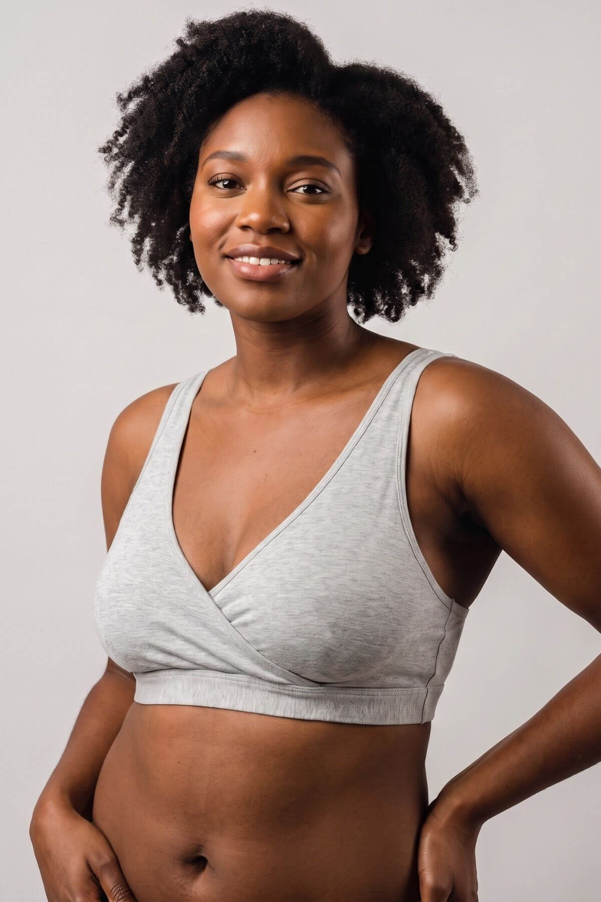 A woman with curly black hair wearing a light gray heather wrap-front bralette, posing against a solid light gray background.