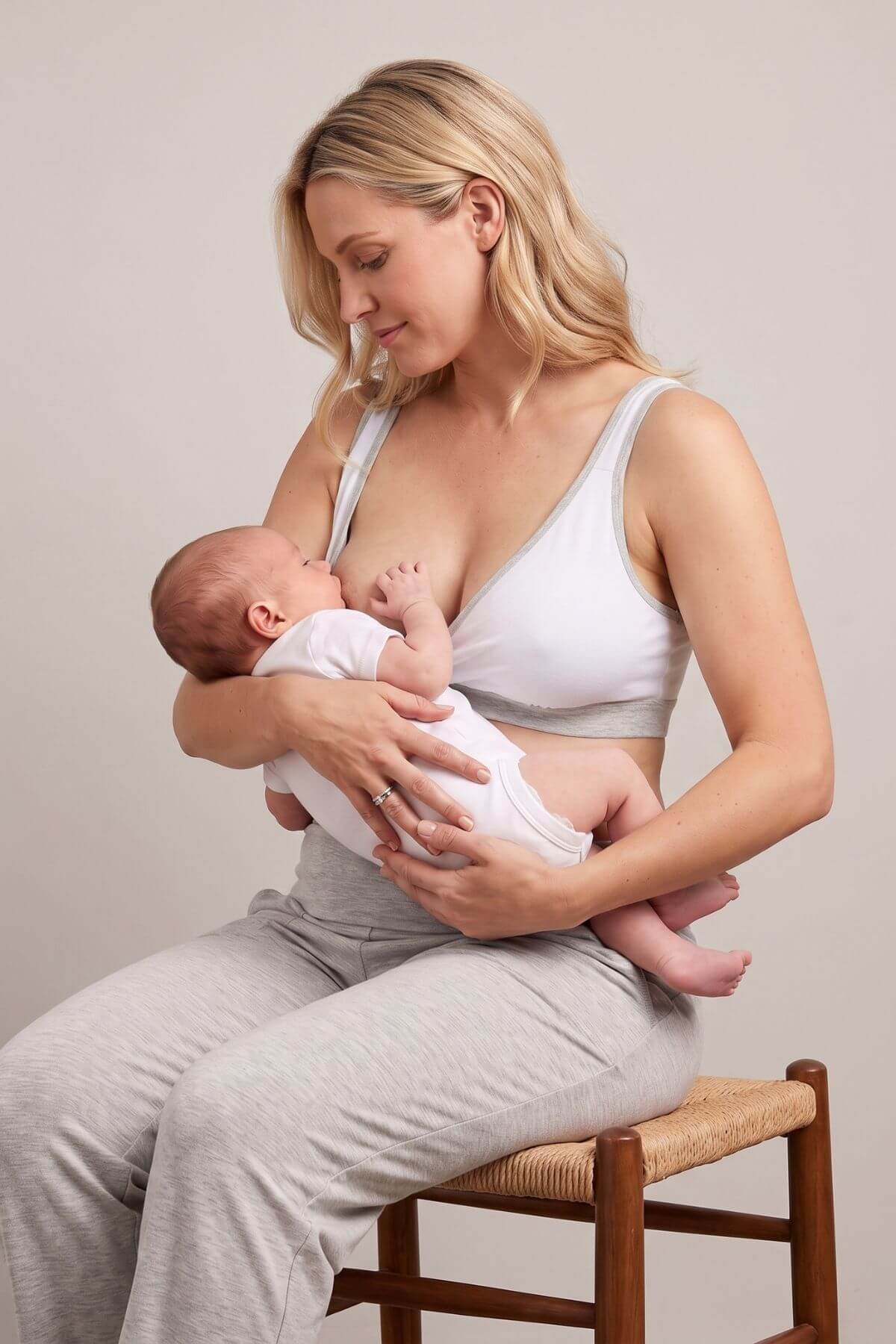 A woman in a white nursing bra and light gray sweatpants sits on a wooden chair, breastfeeding a baby dressed in a white onesie against a plain beige studio background.
