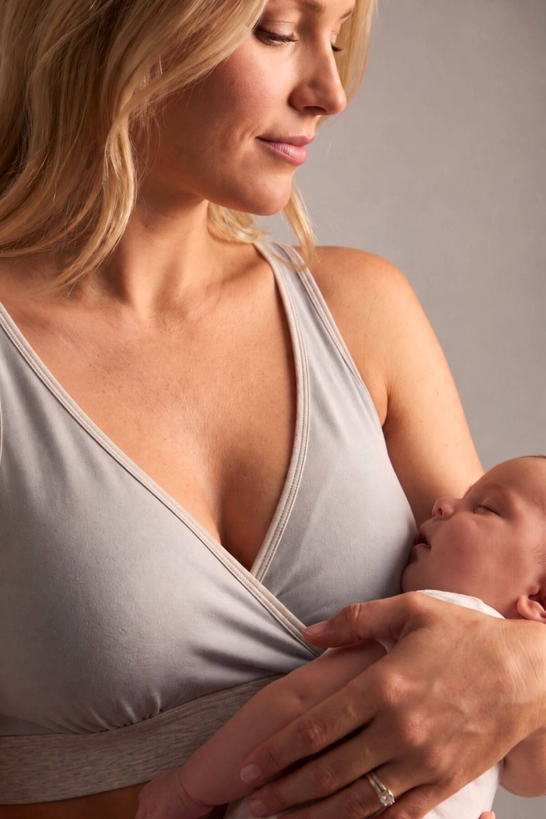 A close-up lifestyle shot of a woman wearing a light gray nursing bra while holding a sleeping newborn baby. The baby is nestled against the soft, wrap-style front of the nursing bra, and the woman wears a silver ring on one of her fingers.