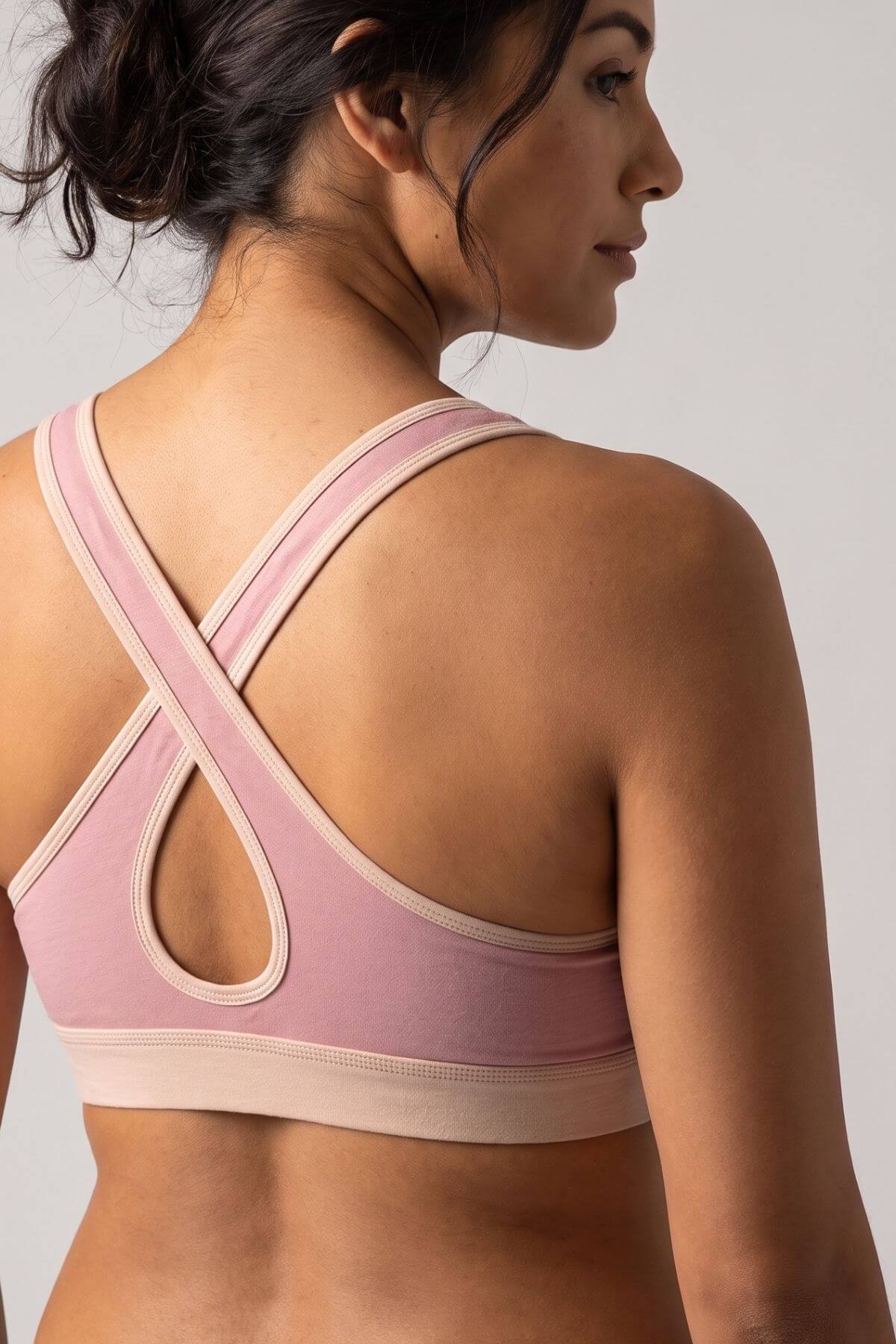 A woman shown from the back wearing a dusty pink sports bra with beige trim and crossover straps, photographed against a light gray studio background.