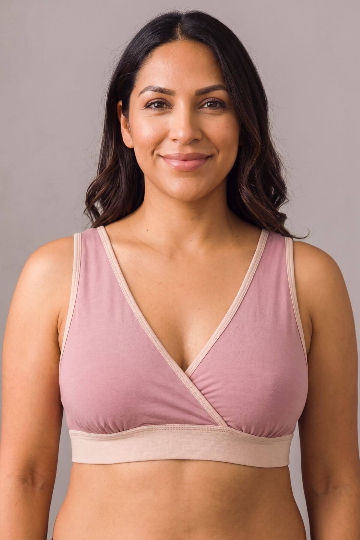 A woman wearing a dusty pink wrap-style bralette with beige trim and a beige underband, shown against a plain gray studio background.