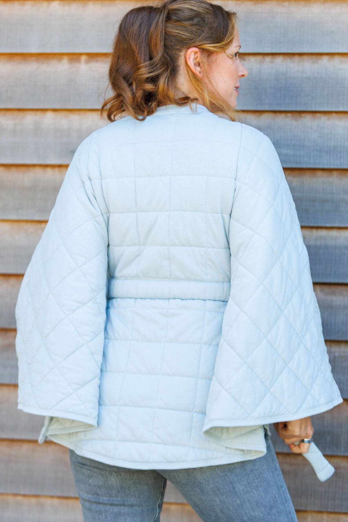 A woman viewed from the back wearing a light blue quilted jacket with wide bell sleeves, standing against a weathered wooden plank wall.