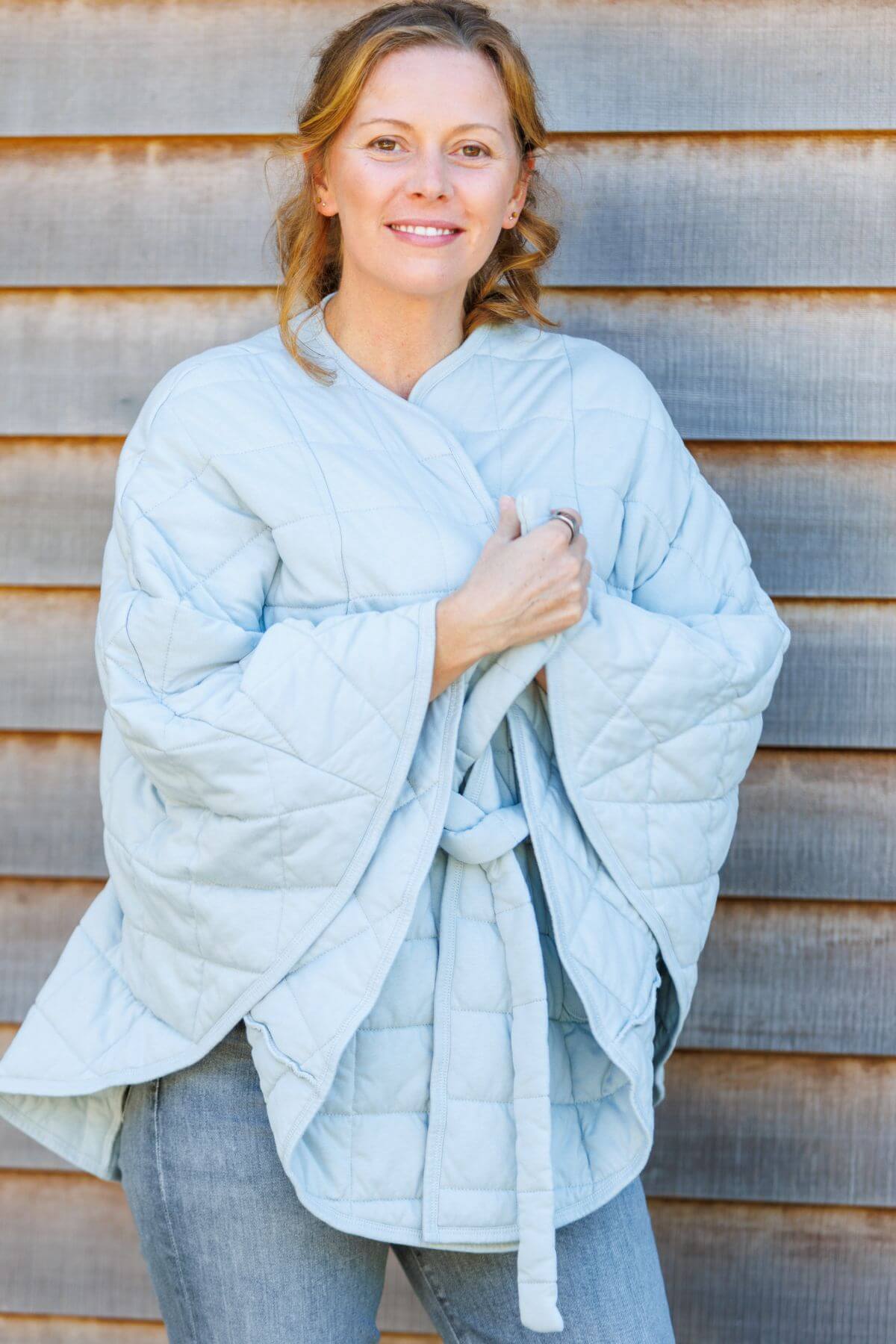 A woman wearing a light blue quilted wrap blanket with a tie belt, standing in front of a rustic wooden plank wall.