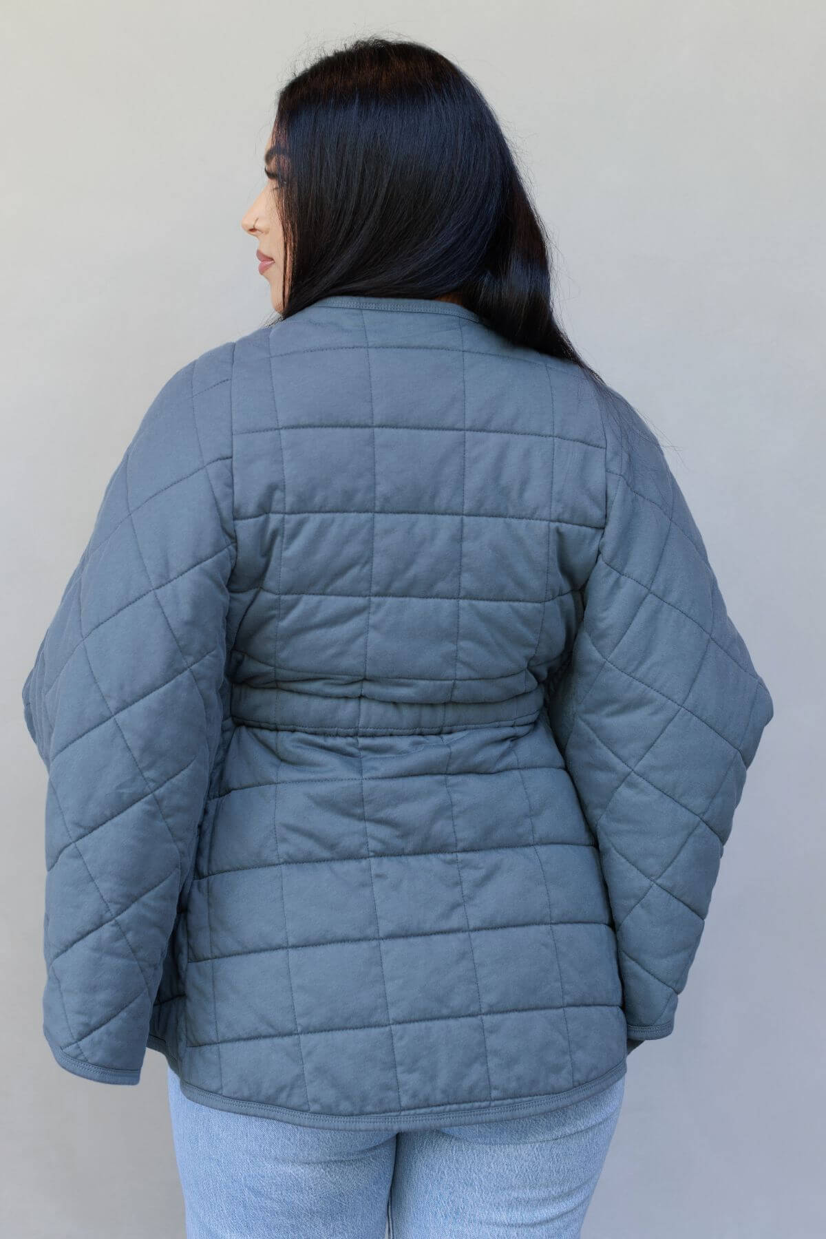 A woman viewed from the back wearing a slate blue quilted puffer jacket and light blue jeans against a plain light gray wall.