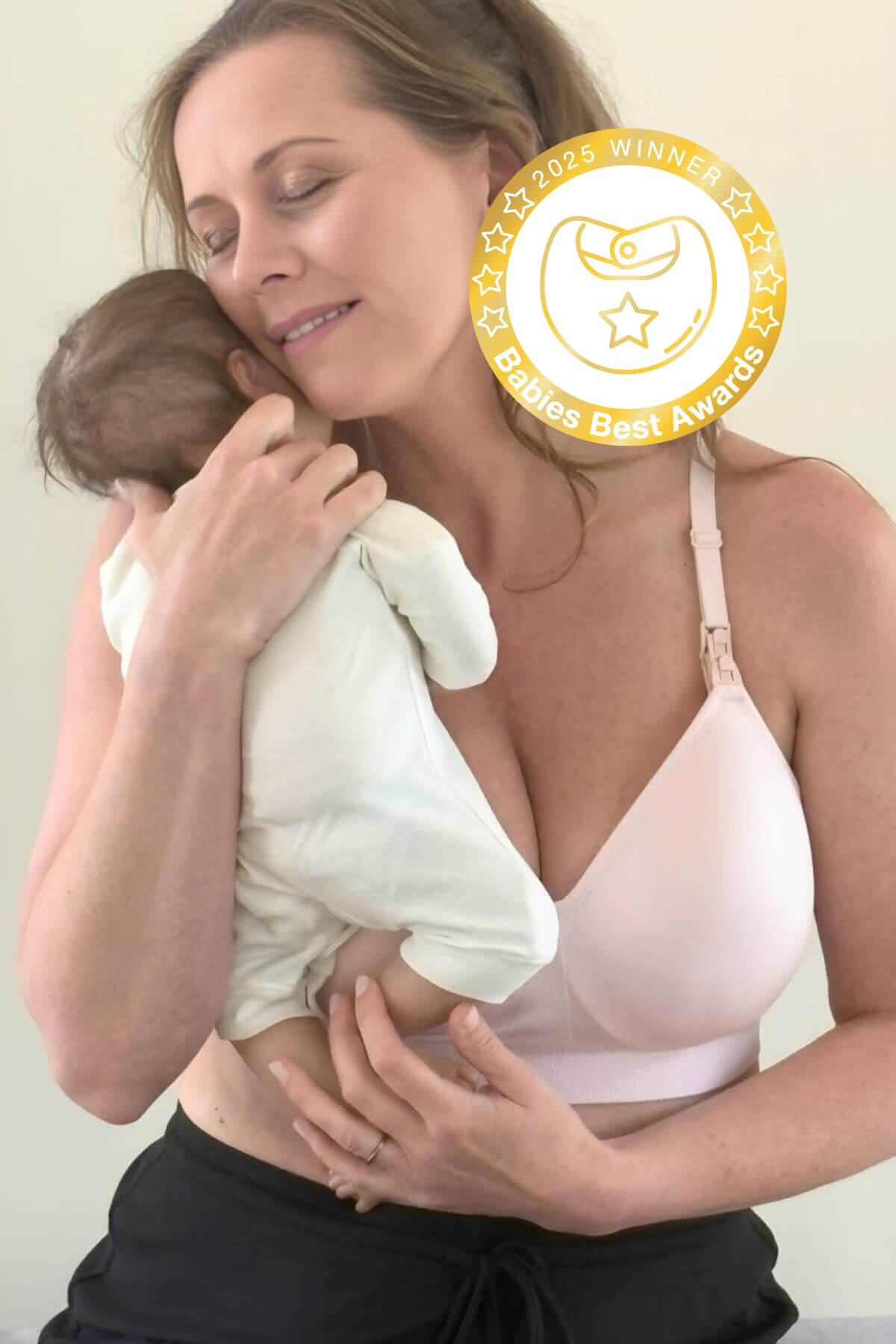 Woman holding a bab wearing a hands free pumping and nursing bra with a badge indicating an award on a plain background