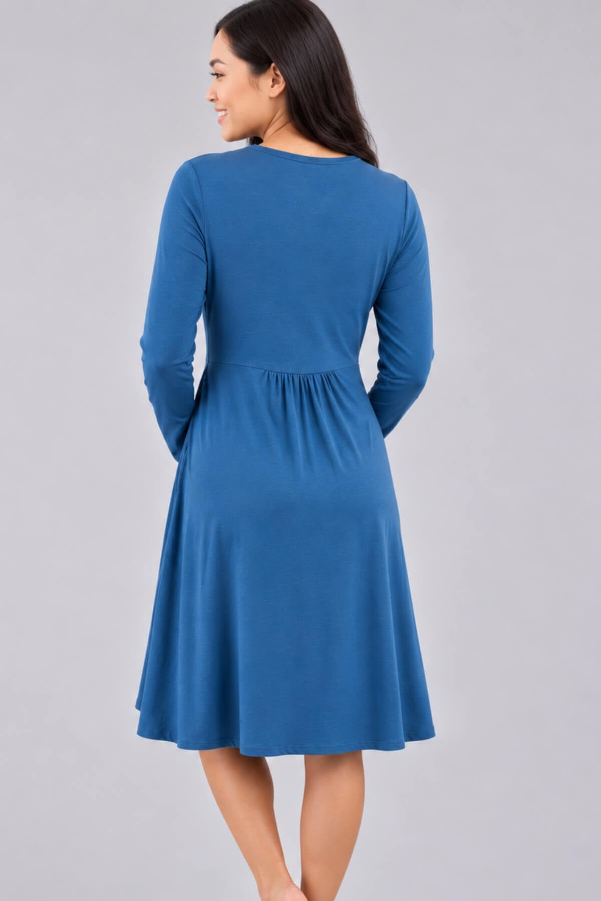 A woman wearing a blue long-sleeve shift dress, shown from the back against a solid gray studio background