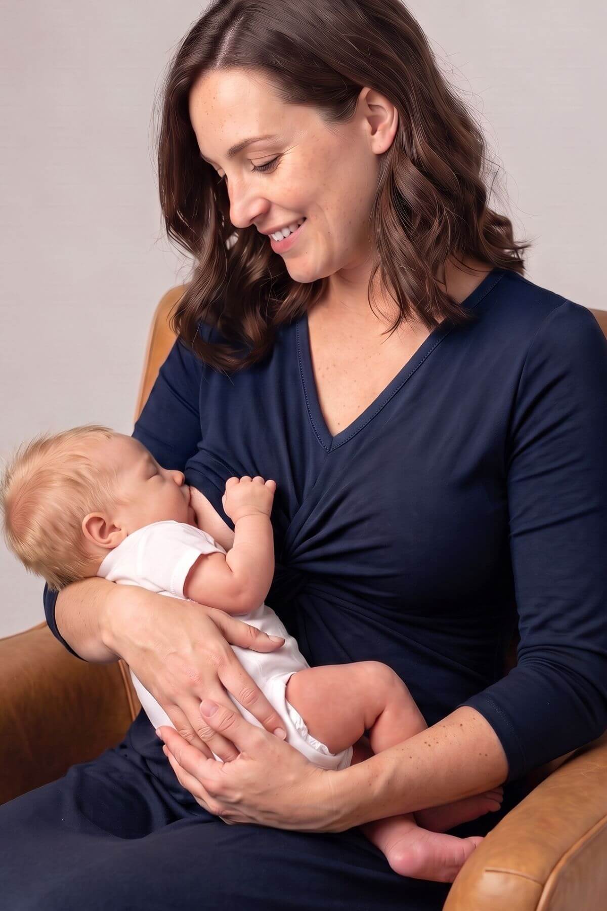 A woman in a navy blue nursing top and matching pants holds a baby wearing a white onesie while breastfeeding, seated in a brown leather armchair against a neutral gray background
